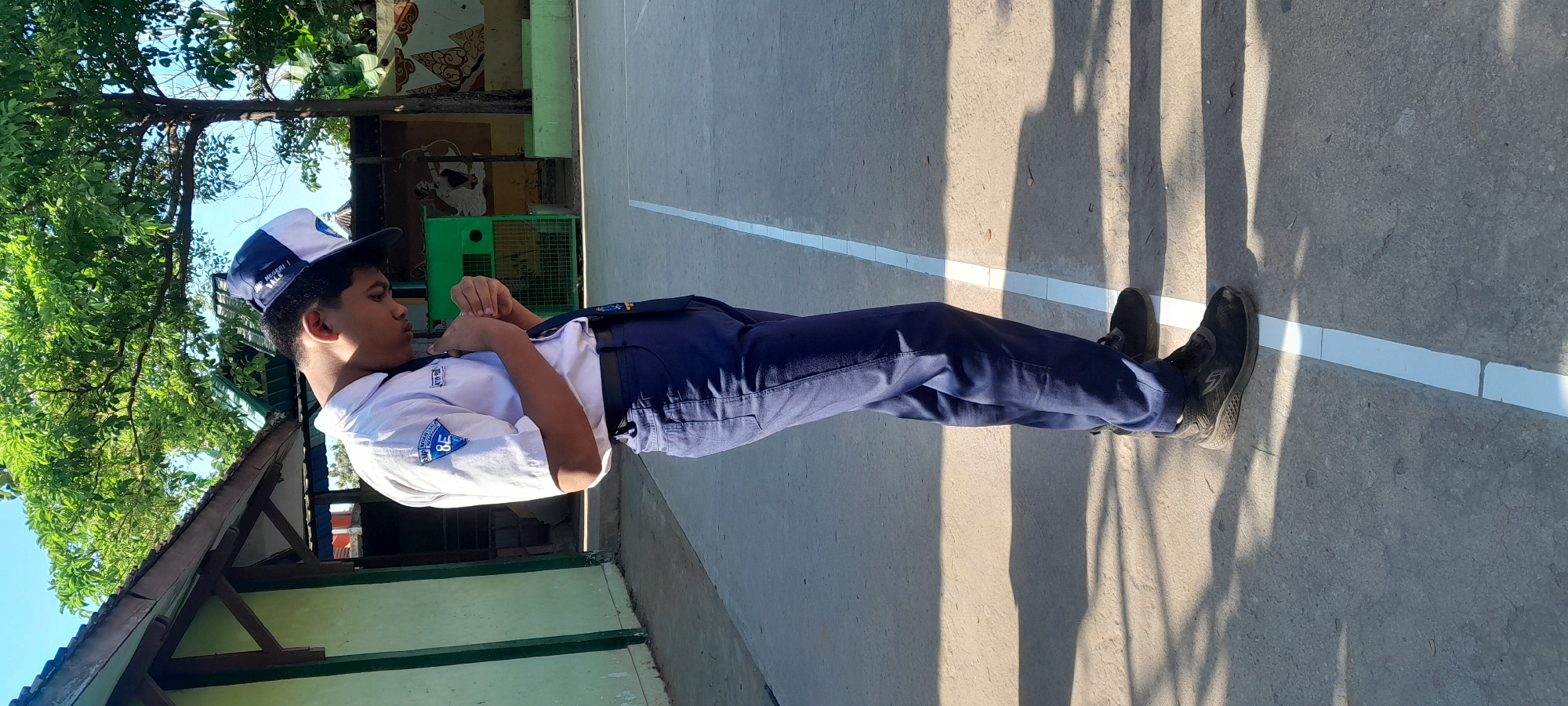 In Tahunan, Indonesia, a young boy in a school uniform stands on an outdoor basketball court. The uniform consists of a white short-sleeved shirt with a blue tie and dark blue pants. He wears a blue and white cap and black athletic shoes. The boy is looking down, with his hands clasped in front of him, as if in prayer or contemplation. The court has a grey asphalt surface with a white line marking its boundary. Behind the boy is a light green building with a wooden roof structure and trees with lush green foliage. Sunlight casts shadows on the court, indicating it is a bright, sunny day.