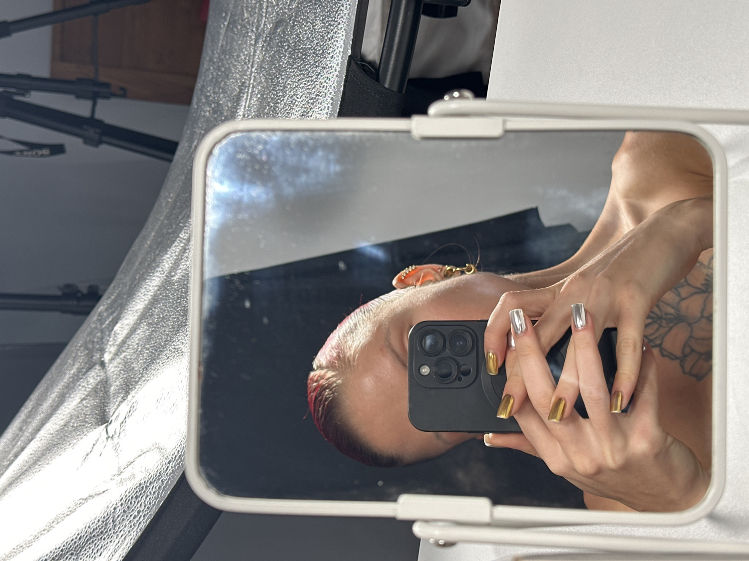 An eye-level mirror selfie captures a person in an indoor setting, likely a photo studio in North Kuta, Indonesia. The person's reflection, oriented with their head tilted to the right, displays short, red or pink-colored hair on top, adorned with a distinctive orange, crab-like ornament. They hold a black smartphone, featuring a multi-lens camera system, in front of their face, obscuring most facial features. Their hands are prominent, showcasing long, square-shaped fingernails painted in an alternating pattern of metallic gold and metallic silver. A visible tattoo with floral or organic motifs extends across their left arm and shoulder area, as seen in the reflection. The rectangular mirror has a simple white frame. In the background, outside the mirror's reflection, a large, silver textured softbox or reflector dominates the left side, indicating a bright, controlled light source. Black components of a light stand or tripod are also visible behind it, with the brand name "SONY" faintly discernable on one part. The scene suggests a professional or semi-professional photography setup.
