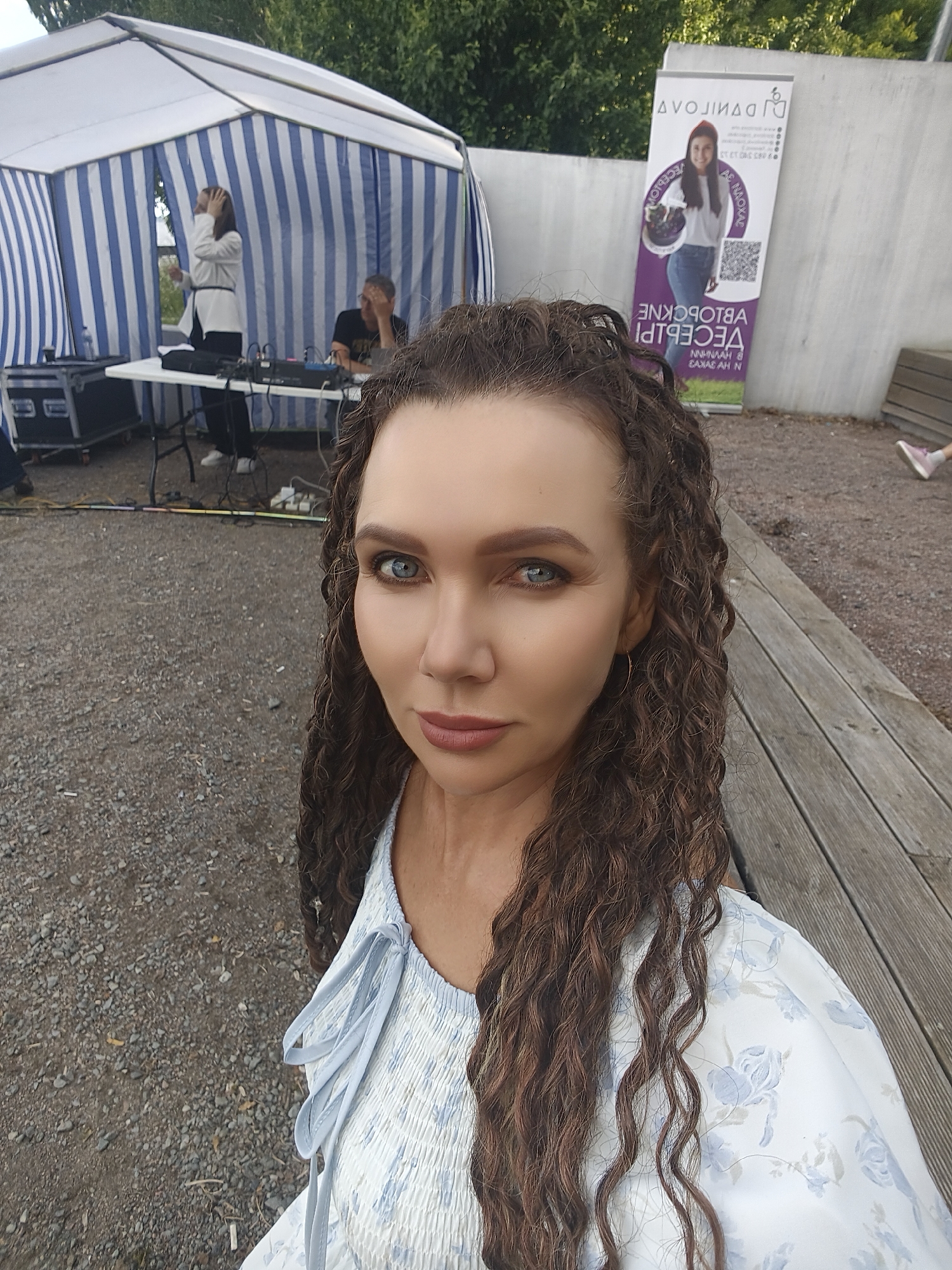This is a selfie taken by a woman with long, curly brown hair and blue eyes, looking directly at the camera. She is wearing a light blue and white floral print top with tie details at the neckline. The background appears to be an outdoor event.
To her left, there is a large tent with blue and white stripes, under which some equipment, including a DJ mixing board and speakers, is set up. A person in a white jacket is standing near the table, and another person is seated behind it.
Further in the background, to the right, a vertical banner is propped against a concrete wall. The banner features text in what appears to be Russian, including a QR code and an image of a smiling woman. Some of the visible text on the banner includes "DJ DANIILOGLI" and "VECEPI".
The ground is covered with gravel, and a wooden deck with benches is visible on the right side of the frame. The lighting suggests it is daytime, with overcast or diffused light. The woman's expression is neutral, and the overall atmosphere seems to be casual and outdoorsy.