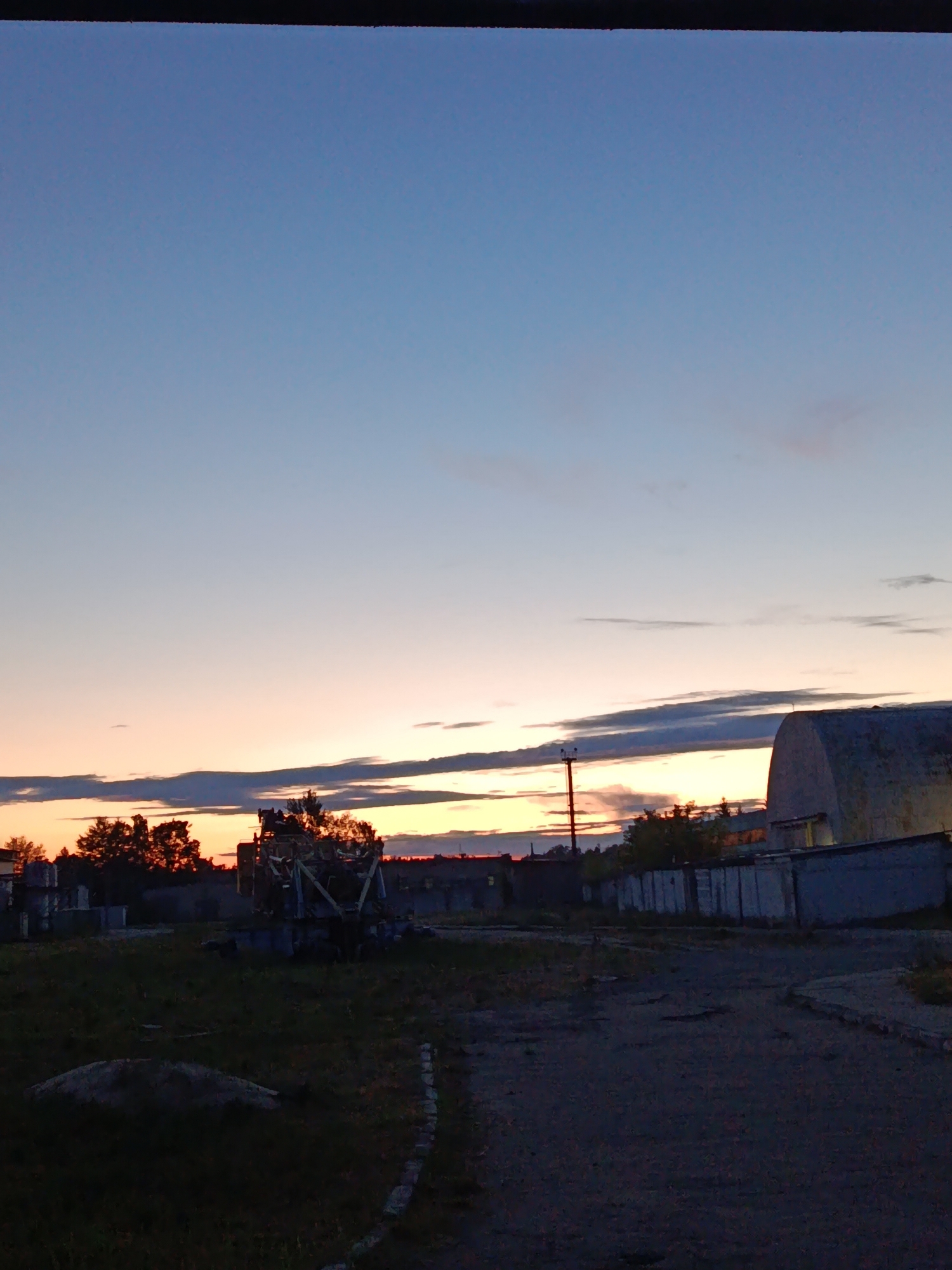 The image depicts a desolate industrial or junkyard area in Svietlahorsk, Belarus, at dusk. The sky is a gradient of pale blue to soft orange and pink, with thin clouds stretched horizontally. In the foreground, a dark, unpaved road or pathway leads into the scene. To the left, there is an area of overgrown grass with a large, indistinct mound of dirt or debris. A large piece of industrial machinery, possibly a crane or construction equipment, is visible in the mid-ground, covered in what appears to be debris or vegetation. Trees with dark foliage are scattered throughout the middle ground, silhouetted against the sky. To the right, several long, low buildings or storage units with corrugated metal walls are lined up. One of these structures, on the far right, is larger and has a curved roof, possibly a hangar or warehouse. A utility pole with a light fixture stands between the road and the buildings. The overall impression is one of neglect and abandonment. There are no people visible in the image.