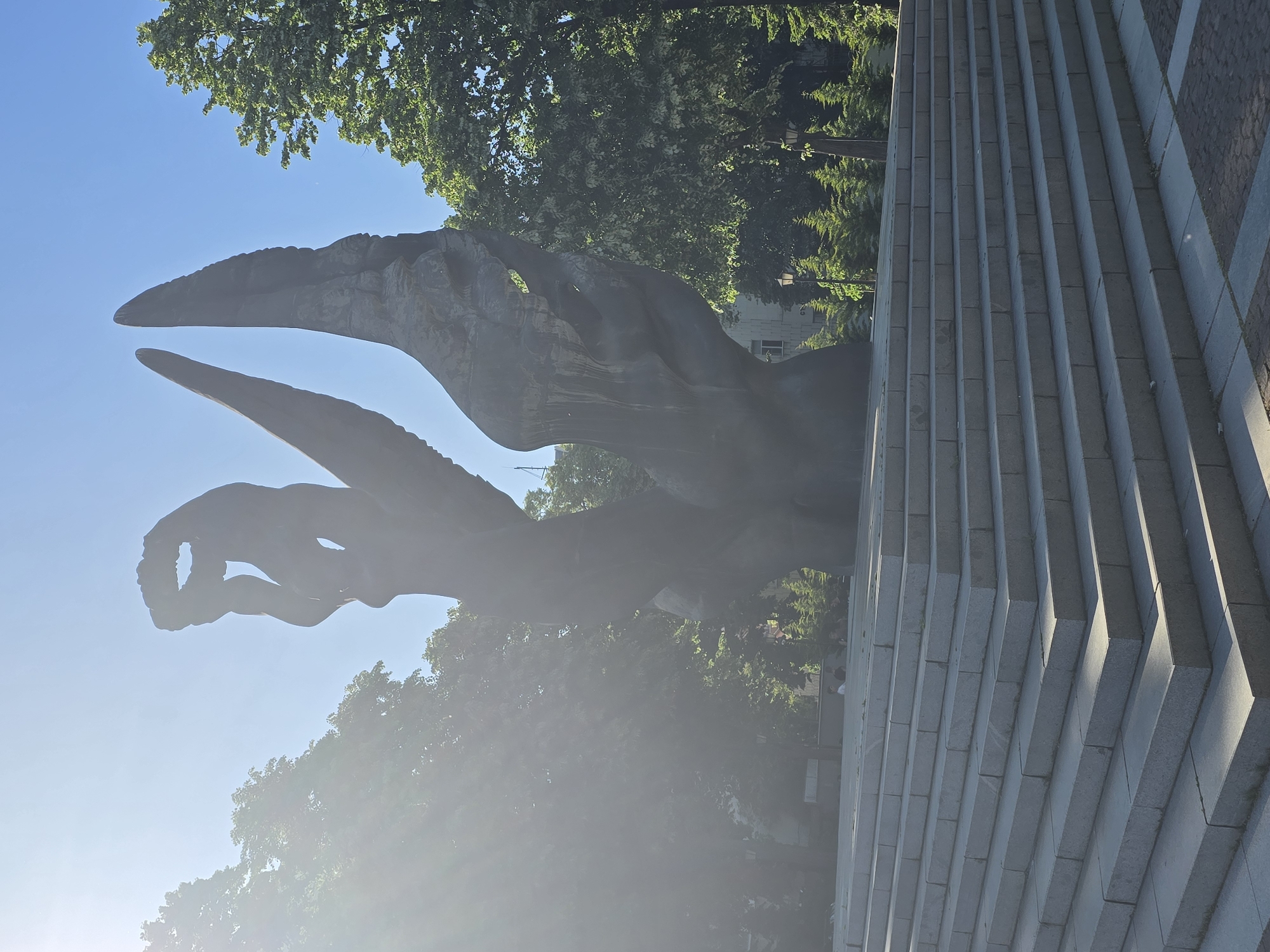 A sideways shot depicts the Monument to the Victims of Fascism, sculpted by Vojin Bakic, in Bjelovar, Croatia. The monument is an abstract, openwork metal sculpture rising against a clear blue sky and surrounded by green trees. To the right of the sculpture is a series of stacked, gray stone blocks or steps. The lower part of the image is somewhat obscured by bright sunlight or mist.