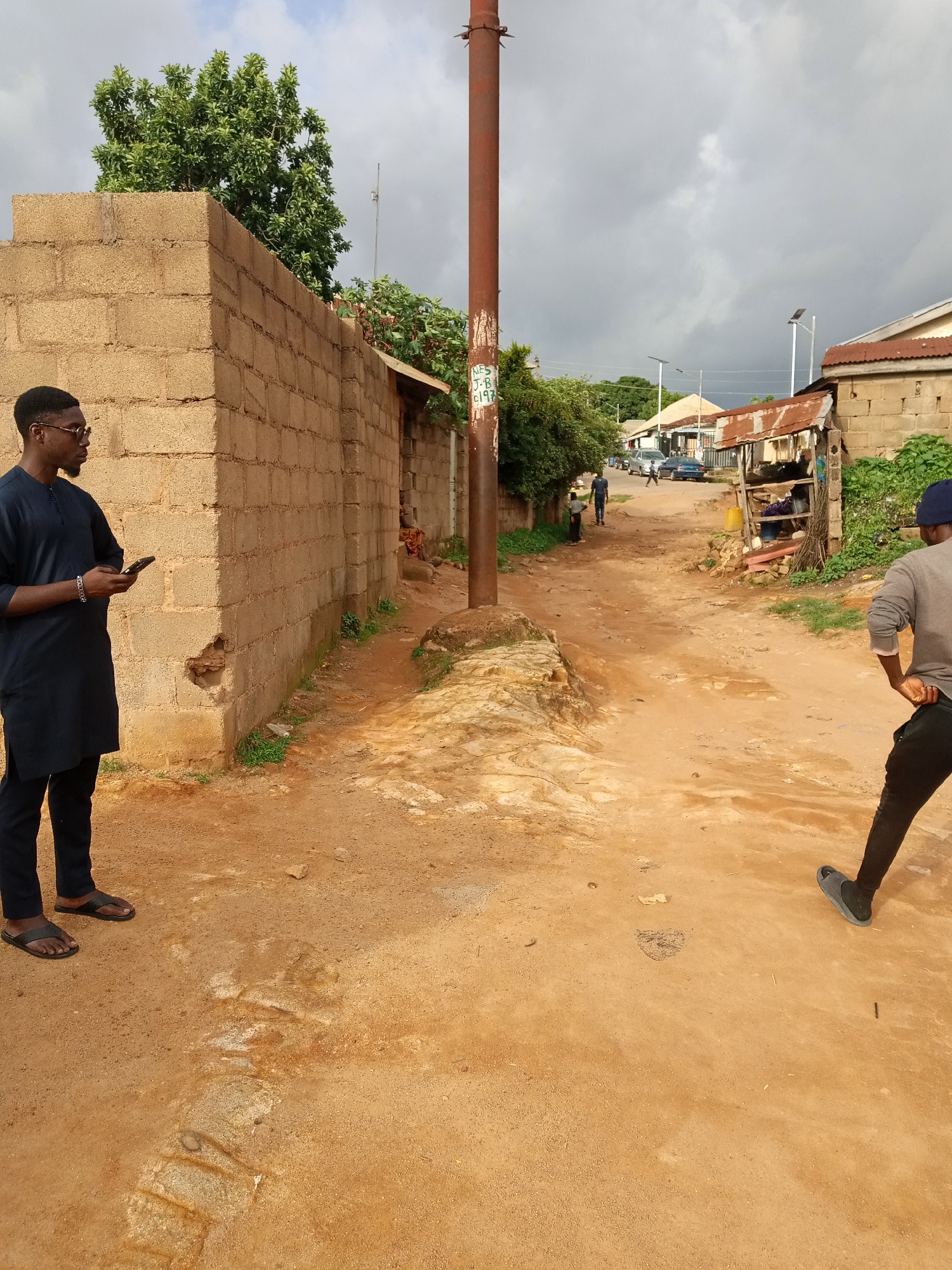 The image depicts a street scene in Jos, Nigeria, under a cloudy sky, suggesting either early morning or late afternoon. The ground is a sandy, reddish-brown dirt road, with some visible rocks and patches of dry vegetation.
In the foreground on the left, a young Black man stands facing left, holding a smartphone in his hands. He is wearing a dark blue tunic with intricate embroidery around the neckline and cuffs, paired with black trousers and black flip-flops. A silver bracelet adorns his left wrist. He appears engaged with his phone.
To the right and further down the road, another person, also Black, is seen from behind, walking away from the camera. They are wearing a gray long-sleeved shirt, black trousers, and a dark blue baseball cap. Their posture suggests they are walking with purpose.
The street is lined with buildings constructed from rough, tan-colored bricks and some structures with corrugated metal roofs, indicating a mix of established and more makeshift dwellings. Trees and lush green foliage can be seen in the background, contrasting with the arid ground. In the middle distance, several cars are parked or driving on the road, and other individuals are faintly visible, suggesting a populated area. The overall scene conveys a sense of everyday life in a developing urban or semi-urban environment. There is some graffiti on a utility pole in the mid-left of the image, but the text is illegible.