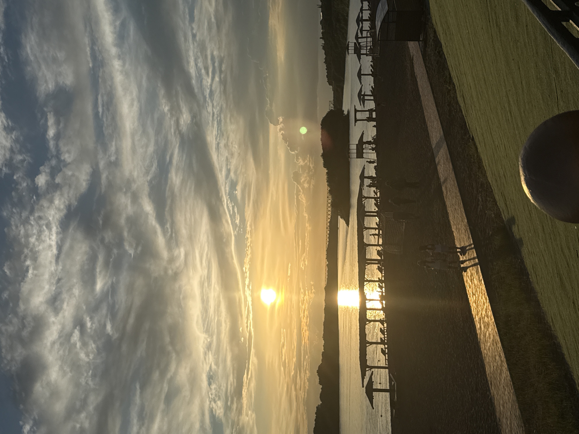 The image depicts a serene sunset over the Irtysh River in Pavlodar, Kazakhstan. The sun, a bright orb, is low on the horizon, casting a golden glow across the water and illuminating the sky with hues of orange and yellow. Wispy clouds, tinged with grey, stretch across the expanse, creating a dramatic backdrop.
On the riverbank, a sandy beach is dotted with numerous shaded cabanas and lounge chairs, suggesting a recreational area. Silhouettes of people can be seen scattered along the beach, some appearing to walk while others are seated. A pathway runs along the sand, bordered by lush green grass on one side and the beach on the other. In the distance, a bridge spans the river, adding a touch of urban infrastructure to the natural landscape. The overall atmosphere is tranquil and picturesque, capturing the beauty of a sunset in this Kazakhstani city.