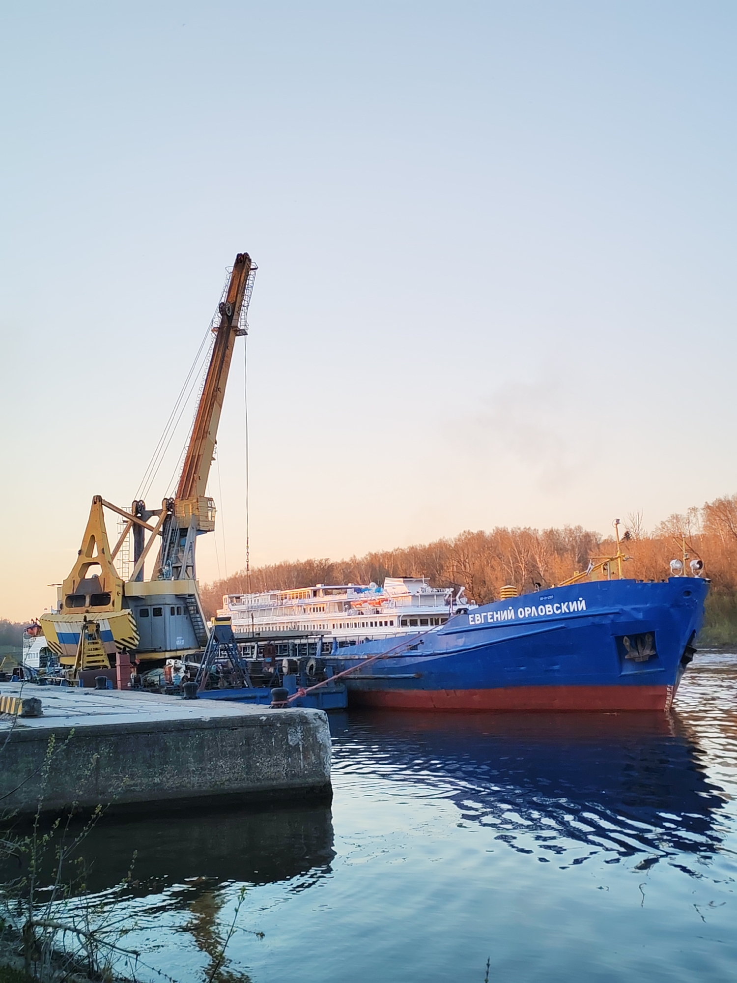 The image depicts a river scene in Yakhroma, Russia. A blue and red ship, labeled "EВГЕНИЙ ОРЛОВСКИЙ," is docked alongside a concrete pier. A yellow and gray crane is positioned on the pier, near the ship. The water shows reflections of the ship and pier. Trees line the riverbank in the background. The sky is light blue and orange, suggesting it's either dawn or dusk. There are no visible people in the primary focus.