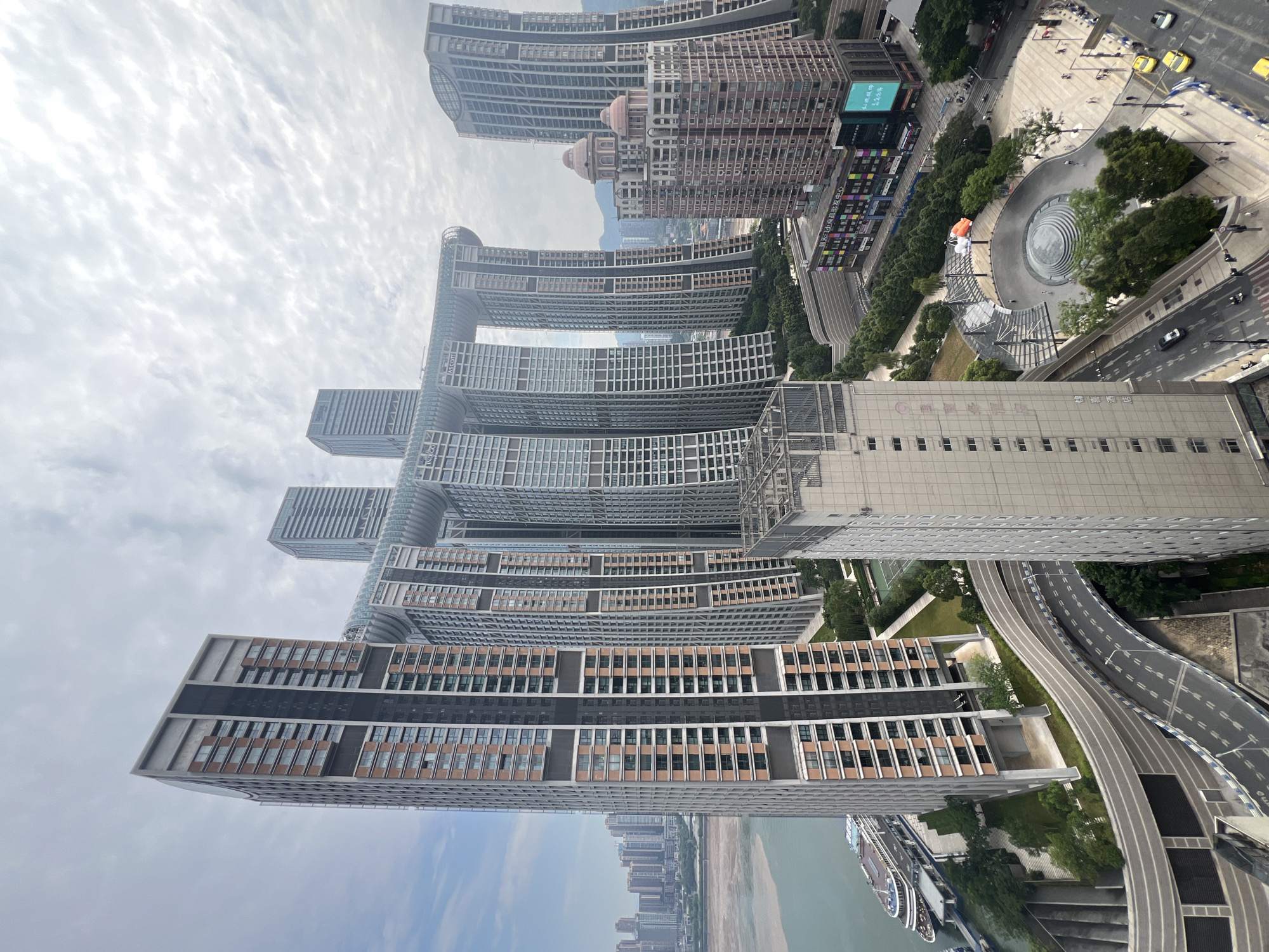 This is a high-angle, medium shot capturing the Raffles City complex in the Yuzhong District of Chongqing, China. The massive, multi-towered structure dominates the frame, with its distinctive horizontal skybridge connecting several of the buildings. The architecture is modern and imposing, characterized by glass and concrete facades. In the foreground, a curved elevated road with multiple lanes is visible, with cars driving on it. To the left, a wide river flows, with a large ferry docked at the waterfront. Green trees and manicured landscaping are interspersed between the buildings and along the riverbank. In the middle ground, a public plaza features a circular water feature and several people walking around. The sky is overcast with dramatic clouds, indicating a potentially humid or impending weather event. The overall scene depicts a bustling, urban environment with a unique architectural landmark.