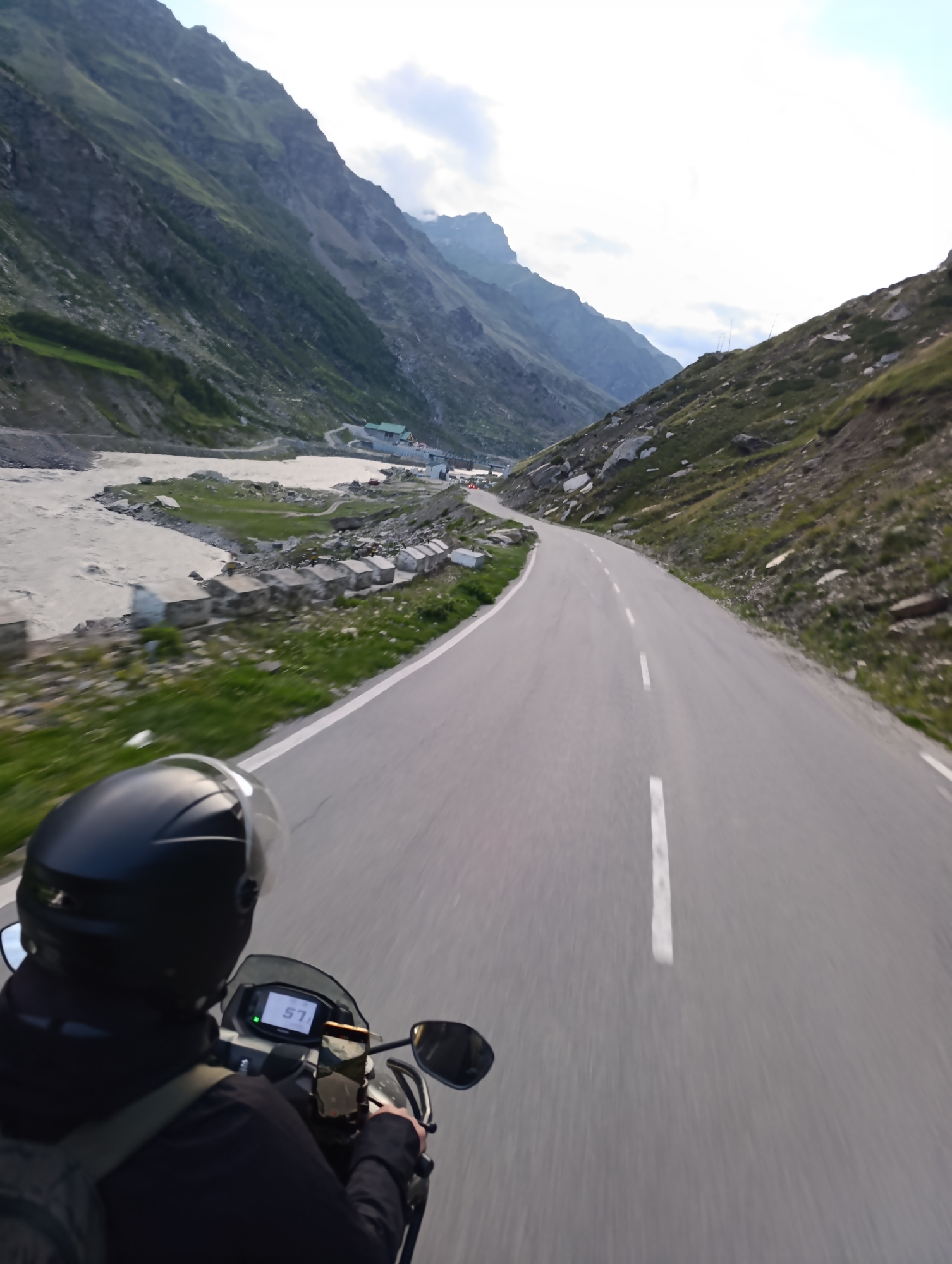 The image shows a first-person perspective of someone riding a motorcycle on a paved road through a mountainous region. The road has a white dashed line marking the center and a white solid line on the right edge. Steep, rocky mountains with patches of green vegetation rise on both sides of the road. To the left, a wide, muddy river flows alongside the road. In the distance, there are some buildings that appear to be part of a settlement or rest stop. The sky is partly cloudy. The motorcycle's dashboard displays a speed of "57". A phone is mounted on the handlebars. The rider is wearing a black helmet and a black jacket. The location is identified as Burwa, India.