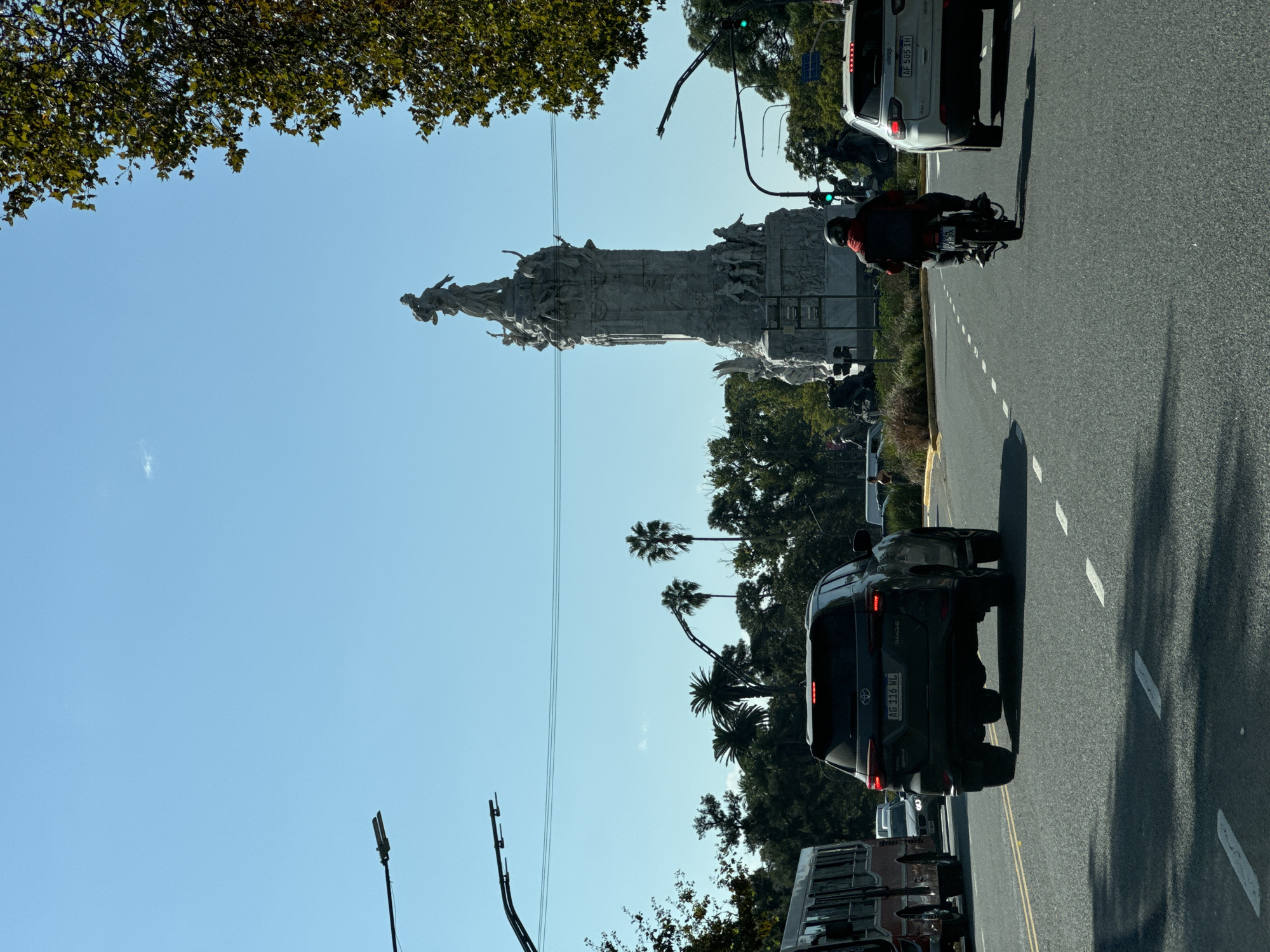 The image shows a street scene in Buenos Aires, Argentina, featuring the Monument to the Carta Magna and Four Regions of Argentina. The monument is a large, ornate structure with numerous statues and figures. The sky is blue, and some trees and palm trees are visible. A motorcycle, a car, and other vehicles drive on the road, marked with white dividing lines.