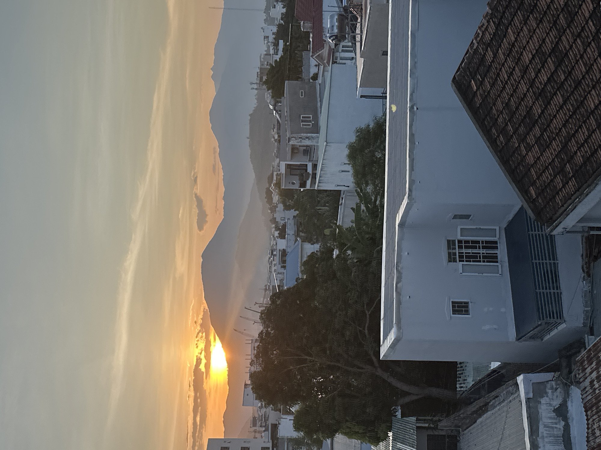 The image depicts a sunrise over Bac Nha Trang Ward, Vietnam, showcasing an urban landscape against a mountainous backdrop. The sun, a bright yellow-orange orb, is partially obscured by a prominent mountain peak on the left horizon, emitting distinct crepuscular rays that fan upwards into the pale blue and orange sky. Scattered wispy clouds are present, particularly in the upper sky.
In the midground, a dense collection of residential and commercial buildings spreads across the terrain, with varying architectural styles. Most structures are multi-story, featuring flat concrete roofs, some with water tanks or solar water heaters visible. Other roofs consist of corrugated metal sheets and reddish-brown tiles. A large, dark green leafy tree stands prominently in the lower-mid left, casting a shadow. Beyond the immediate buildings, the city extends towards distant, hazy blue-grey mountain ranges that define the horizon line. A slender, dark communication or utility tower is visible on the far right horizon, silhouetted against the sky.
The foreground is dominated by the roofs and upper facades of nearby buildings. A large, light-grey building with a flat roof, featuring a small square window and a larger rectangular window with open white shutters, is prominent in the bottom-mid section. Below its large window, a horizontal slatted balcony or screen is visible. To the bottom right, a segment of a tiled, dark brown roof is present. No people or overt human activity are visible within the frame.