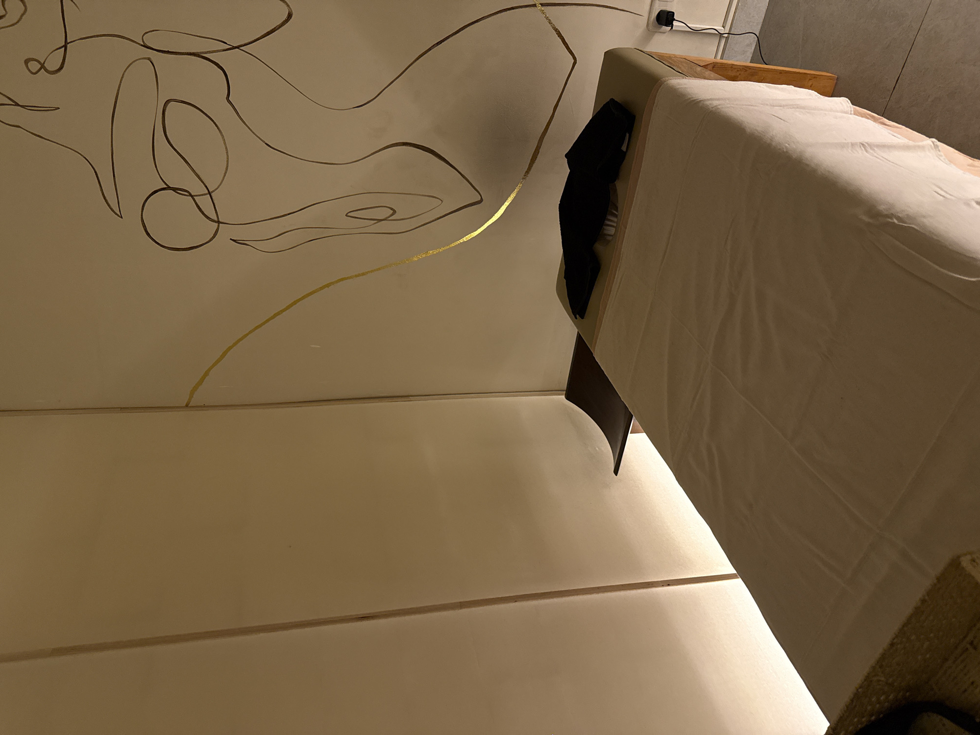 The image captures an interior room, likely a bedroom, from a low, upward-angled perspective. The main subject is a light beige wall adorned with abstract line art. The artwork features flowing, continuous lines rendered in a dark brown color, creating an organic, minimalist pattern. A prominent, shimmering gold line also cuts across the wall, interacting with the brown lines.
To the right, part of a bed is visible. It is covered with a light-colored, possibly white or off-white, sheet or duvet. The bed's headboard appears to be upholstered in a light beige or tan fabric, with a wooden element behind it. A dark, possibly black, item of clothing is draped over the headboard. Above the bed, on the wall, an electrical outlet with a black power cord plugged into it is visible.
The lower portion of the image shows a different section of the room, possibly a lower wall or floor, composed of light-toned horizontal panels or sections. The lighting throughout the scene is warm and appears to be artificial, suggesting an indoor setting, likely in the evening or at night. No people are present, and there is no visible text. The overall impression is one of a decorated, private space.