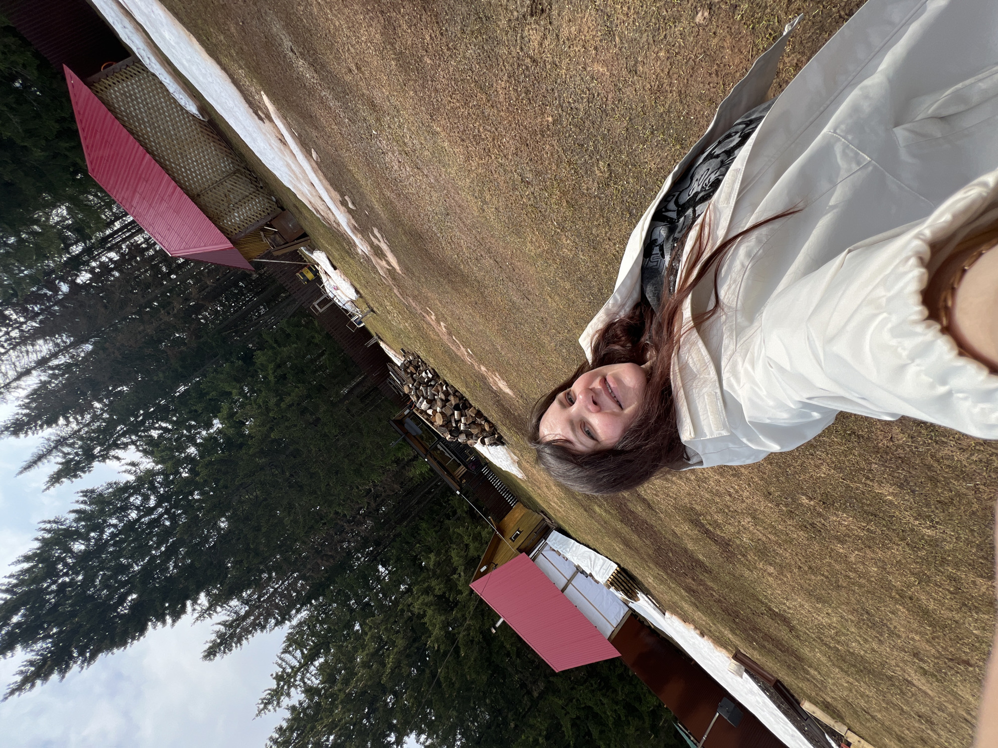 An outdoor portrait captures a young woman smiling directly at the camera, positioned on the right side of the frame. She has long, dark brown hair and is wearing a light-colored, possibly white or light gray, jacket over a darker, patterned shirt. Her expression is joyful and relaxed.
The scene depicts a rural or semi-rural landscape in Bol'shakino, Russia, characterized by patches of dry, brown grass and melting snow, suggesting late winter or early spring conditions. Tall, dark green coniferous trees line the left side of the background, reaching up towards an overcast sky. In the mid-ground, several small buildings or structures are visible, including one prominent structure with a distinctive red, angular roof. A stack of firewood is also present, hinting at the colder climate. The lighting is bright and diffuse, indicating daytime. No visible text is present in the image.