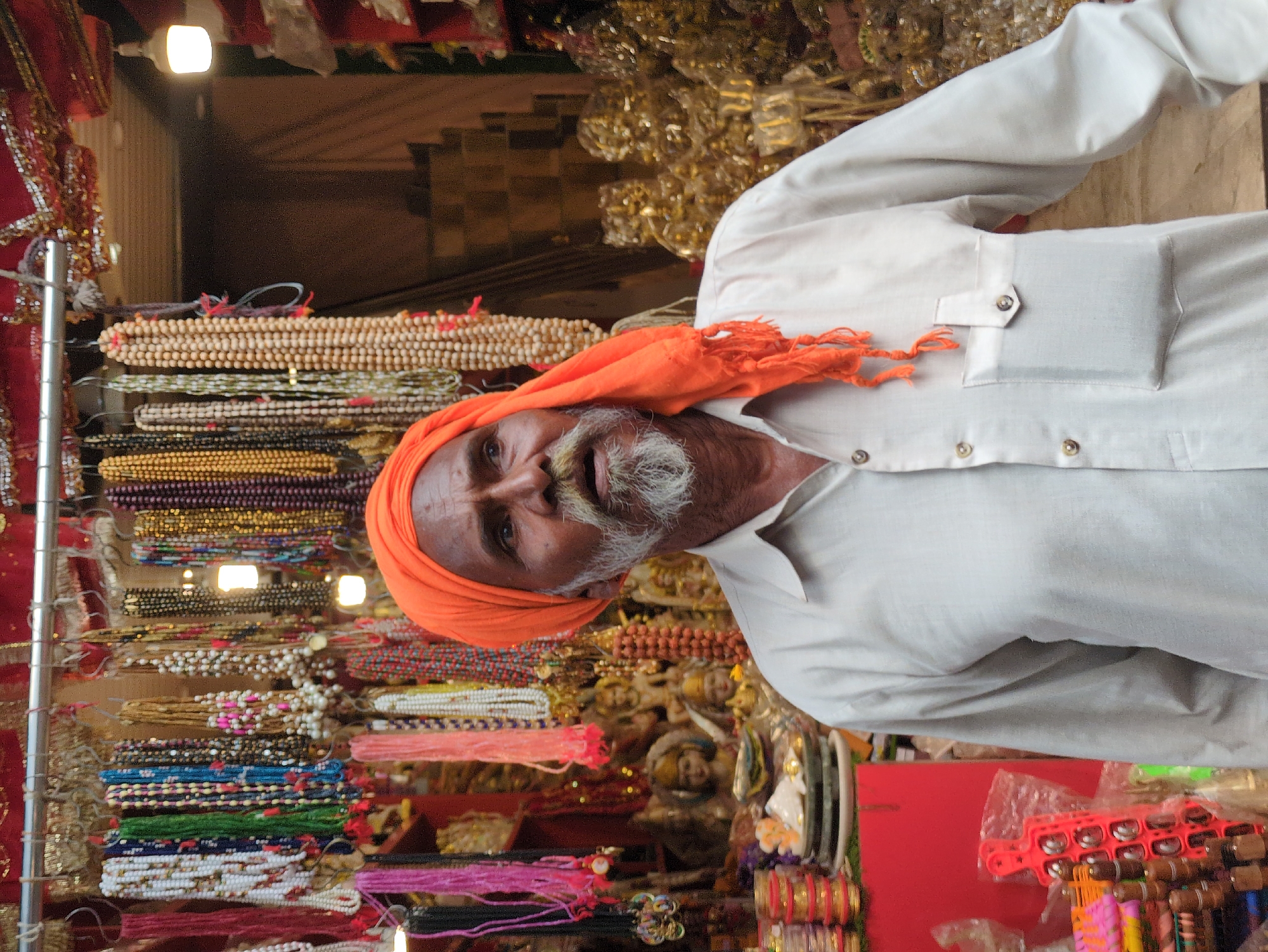 The image shows a medium shot of an elderly South Asian man wearing an orange turban and a light-colored shirt. He has a grey beard and mustache. He is standing in front of a stall filled with religious items, likely in a marketplace. The stall displays numerous strings of beads, some made of wood and others of colorful stones or pearls. There are also small statues and other trinkets visible. The background is somewhat blurred, but suggests a bustling environment with other stalls or shops. The lighting appears to be artificial, possibly from bulbs within the stall. The man is looking to his right, with a neutral expression. This scene is likely from Kangra, India, given the religious nature of the items and the presence of a man in traditional attire.