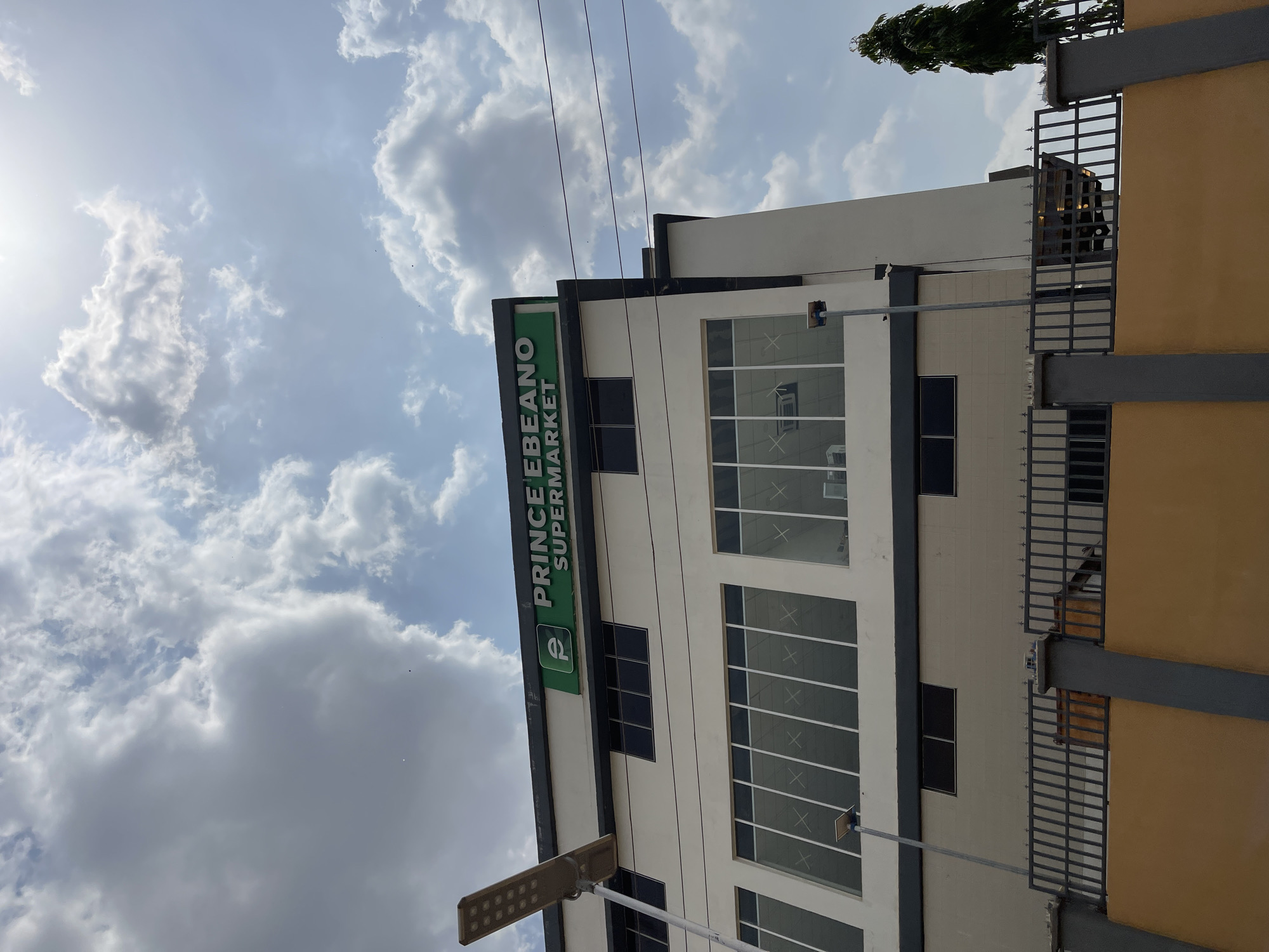 The image displays an upward view of a modern commercial building under a bright, partly cloudy sky in Abuja, Nigeria. The building features a clean, off-white facade with dark trim, contrasting with numerous large windows that appear to have a grid-like or X-shaped internal structure. A prominent rectangular green sign is affixed to the building, bearing the text "P PRINCE EBEANO SUPERMARKET." The 'P' is integrated into a distinctive logo within a green square.
To the left, a contemporary streetlight equipped with multiple LED panels extends upwards into the frame. Above, thin power lines or cables are visible against the light blue sky, which is filled with scattered white cumulus clouds. A bright sun glare emanates from the upper left quadrant, indicating a sunny daytime scene. On the right side, dark grey metal railings suggest a boundary or balcony of an adjacent structure, next to a wall painted in a warm yellow-orange color. A portion of a tall, slender green tree is visible at the extreme top right. No people or specific activities are depicted; the scene focuses on the architectural elements and the sky.