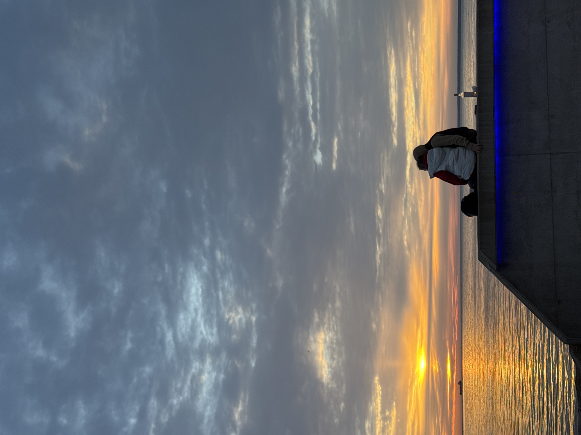 In Tallinn, Estonia, two individuals are silhouetted against a twilight sky as they sit on a concrete ledge overlooking the sea. The sky is filled with dramatic clouds, with hues of gray, blue, and hints of orange from the setting sun. The sun's rays are reflecting off the calm water, creating a golden shimmer. In the distance, a ship is visible on the horizon. The couple is seated close together, suggesting an intimate moment. A bottle stands upright on the ledge next to them. Blue lights illuminate the underside of the ledge. The scene evokes a sense of peace and romance.