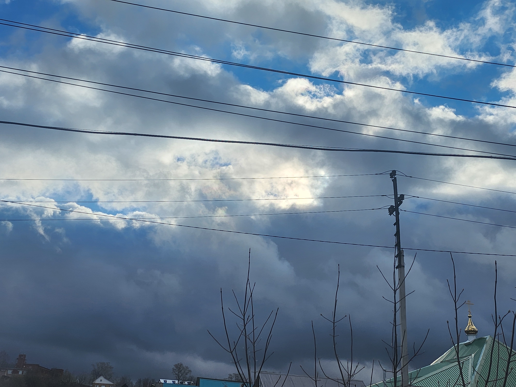 This daytime image captures a dynamic sky over Krasnogorskoye, Russia, characterized by a mix of bright white cumulus clouds and darker, heavy grey clouds, interspersed with clear patches of blue sky. Numerous black power lines crisscross the upper and middle sections of the frame. Sunlight penetrates through a luminous opening in the clouds in the mid-left, creating a bright spot.
In the foreground and mid-ground, the bare, leafless branches of deciduous trees rise towards the sky, suggesting late winter or early spring. A prominent grey utility pole stands on the right side, supporting several power lines. Below the cloud line, a building with a green, corrugated metal roof is partially visible on the right, distinguished by a golden, onion-shaped dome topped with a cross, indicating an Orthodox church. On the far left, indistinct buildings are visible on a distant hillside. No people are present, and no text is visible. The scene conveys unsettled weather conditions under a dramatic sky.