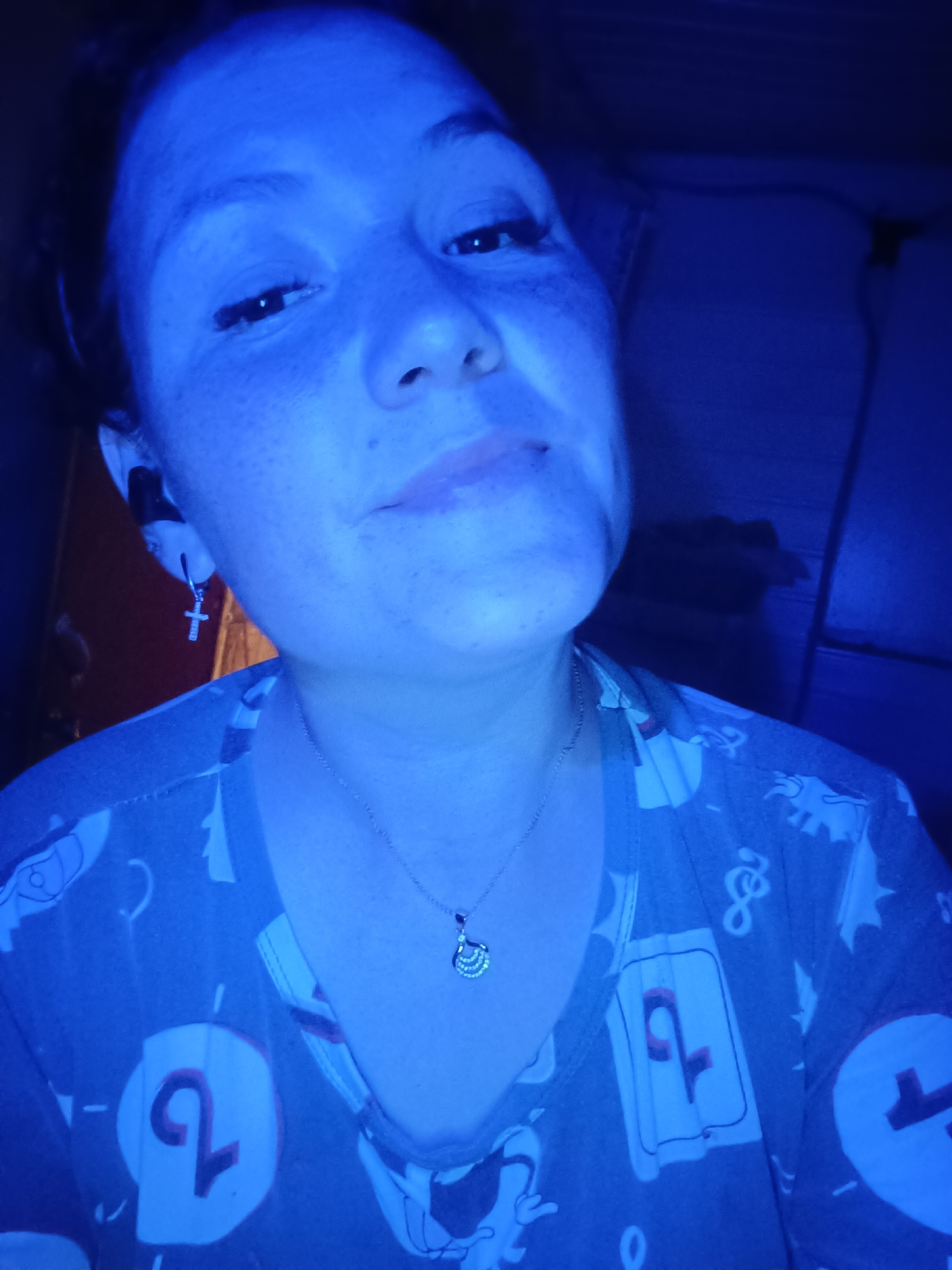 In this close-up, dimly lit, blue-toned selfie, a person's face and upper torso are visible. The individual has dark hair pulled back, and their face is angled slightly to the right. They are wearing a grey shirt with a pattern of white shapes, possibly cartoon characters or symbols, and some red accents, including what appears to be the number "2" on a white card-like background. A delicate silver necklace with a small, ornate pendant rests on their chest. A silver cross earring is visible on their left ear. The lighting casts a strong blue hue over the entire scene, obscuring most details of the environment, which appears to be indoors.