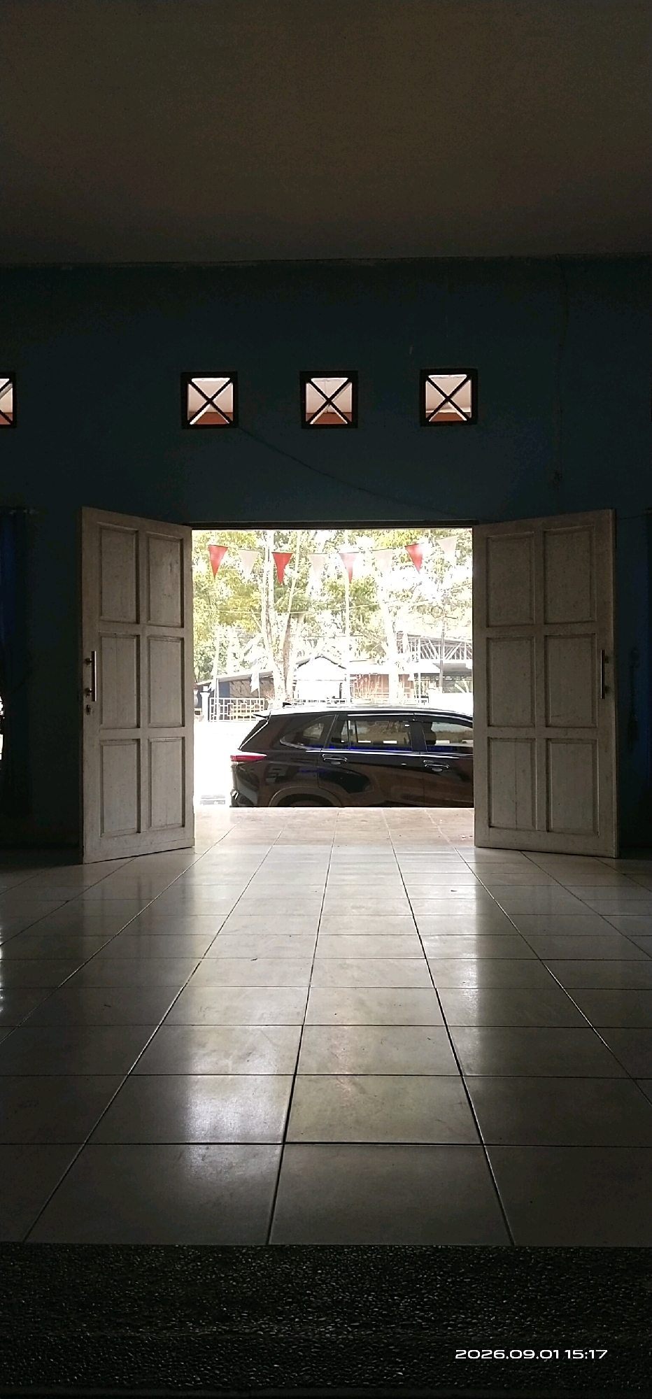 The image captures a view from inside a building, looking out through an open doorway. The interior features a tiled floor that reflects the light, and a teal-colored wall with three square windows, each containing an "X" shaped grate, spaced evenly across the top portion of the wall. The doorway is framed by two large, wooden, paneled doors, one of which is open.
Outside, a dark-colored SUV is parked, partially visible through the doorway. Beyond the vehicle, trees and some structures suggest an outdoor environment, possibly a street or a courtyard. Red and white triangular flags are strung across the scene outside, suggesting a festive or celebratory occasion. The overall lighting indicates it is daytime, with a strong light source coming from outside. A timestamp in the lower right corner reads "2026.09.01 15:17". The location is Tamiang Layang, Indonesia.