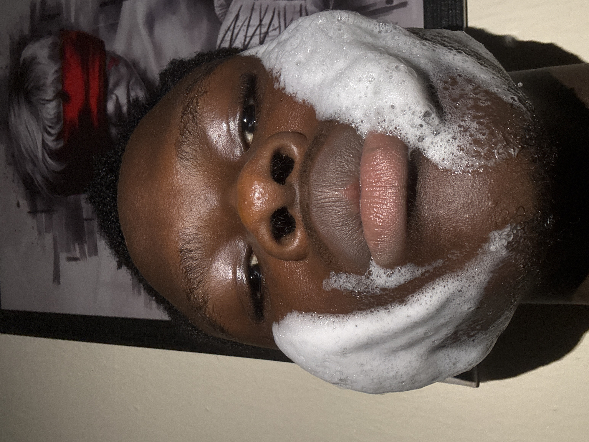 The image displays a close-up, vertical shot of a person's face from the chin to just above the eyebrows. The individual has dark skin and short, dark, curly hair visible at the hairline. White, frothy foam, consistent with soap or a facial cleanser, heavily covers both cheeks and extends down to the lower jawline. The person's lips are slightly parted, revealing a textured surface. The nose is prominent, with visible nostrils. The eyes are open and dark, looking directly at the viewer, with distinct reflections indicating a direct light source. The forehead shows a slight sheen.
In the upper right background, a framed picture is visible. The image within the frame is grayscale and depicts a person, possibly a child or young adult, with light-colored hair or a wig, wearing a vivid red blindfold across their eyes. Red markings are also present on the lower part of the figure's face in the picture. The framed picture has a dark border. To the left of the framed picture and the person's head, a plain, light-colored wall is visible. A shadow is cast onto the lower left wall by the person's head. No specific geographical location, such as Ihiagwa, Nigeria, is discernible from the visual content of the image.
