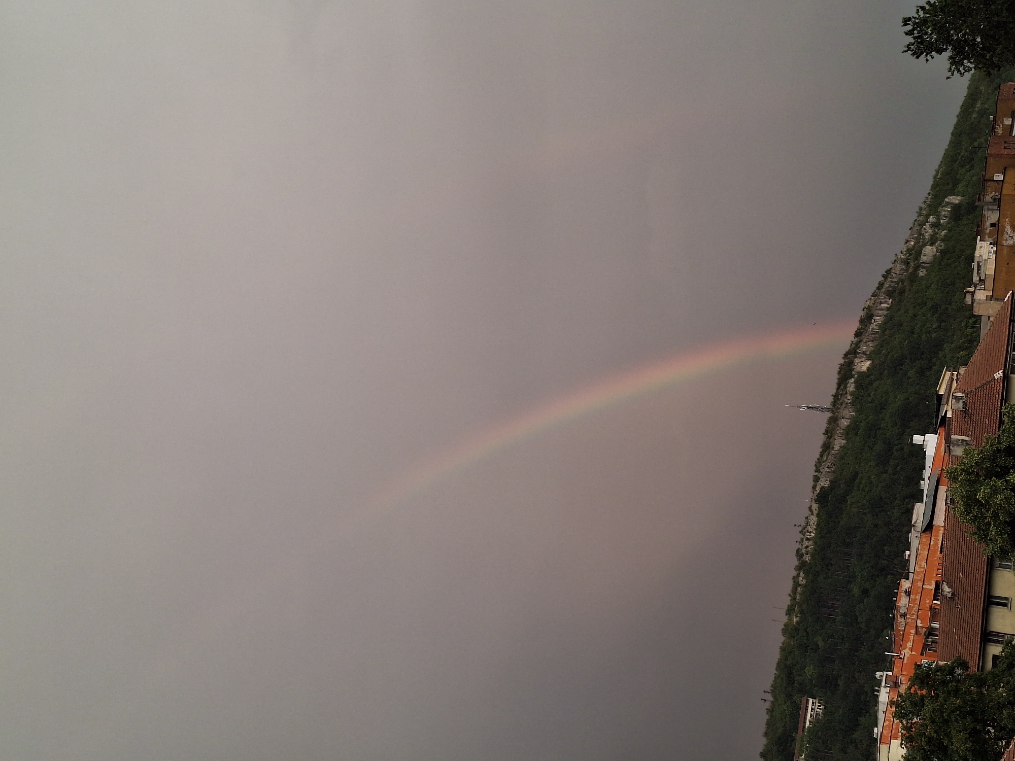 This image depicts a panoramic view of the city of Lovech, Bulgaria, under a cloudy sky with a rainbow arching across it. The foreground features a collection of buildings with terracotta tile roofs, some displaying orange and white facades. Trees are scattered throughout the urban landscape, and a dense forest covers the slopes of a hill in the mid-ground. A rocky outcrop with sparse vegetation is visible on the hillside, along with what appears to be a communication tower. The sky is overcast, with the primary rainbow prominently displayed, and a fainter secondary rainbow is also discernible. The overall atmosphere is that of a post-rainy or pre-stormy weather event.