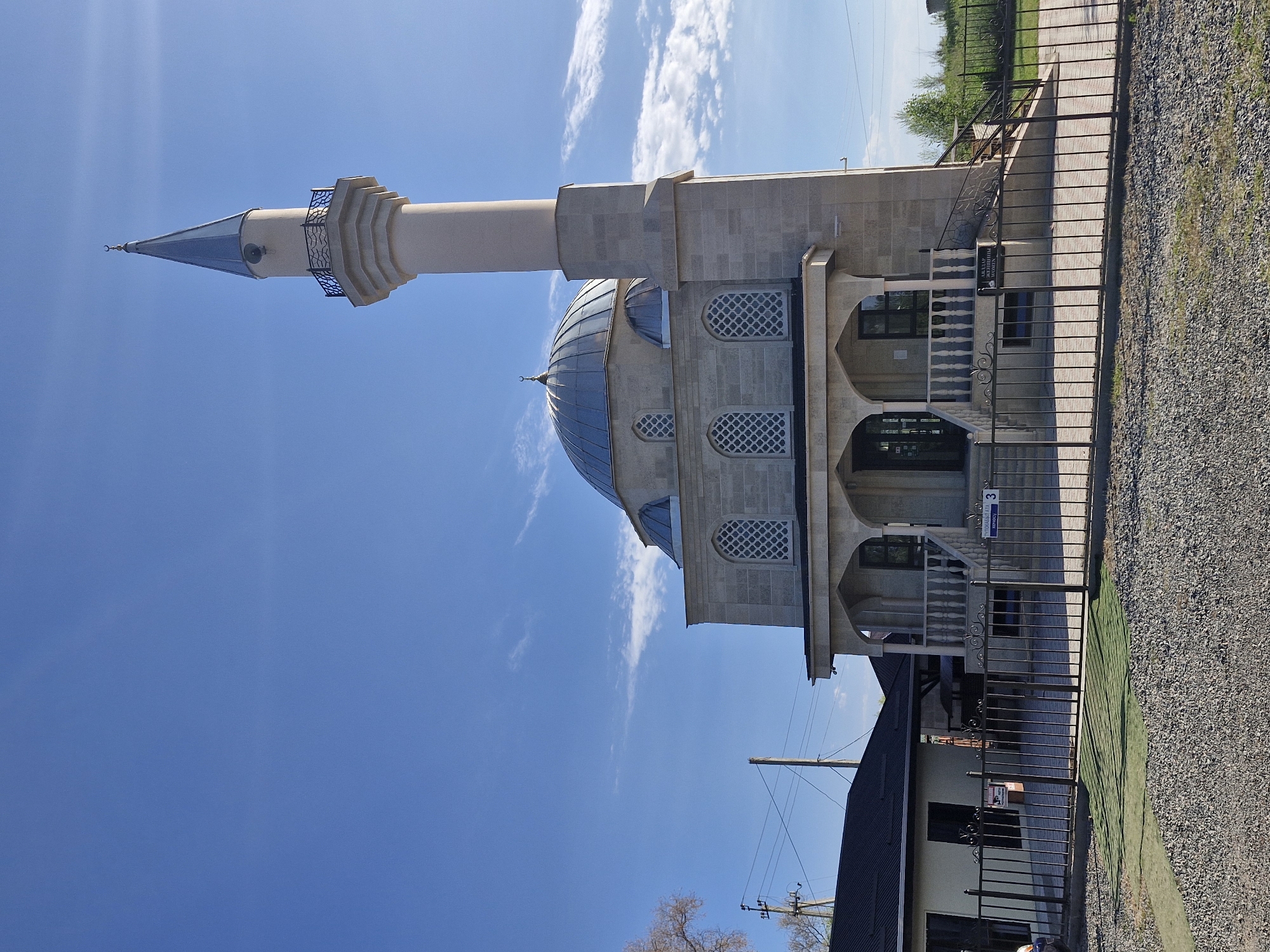 In the city of Bulan-Segettu, Kyrgyzstan, a mosque with a light-colored stone facade stands under a bright blue sky with scattered white clouds. The mosque features a prominent dome and a tall, slender minaret topped with a crescent moon. An arched entrance leads to the building, flanked by ornate railings and steps. Adjacent to the mosque is a smaller, dark-roofed building. A black metal fence encloses the property, which is surfaced with gravel and some patchy green turf. Power lines and utility poles are visible in the background, along with some trees and vegetation. The scene is static and depicts a clear day.