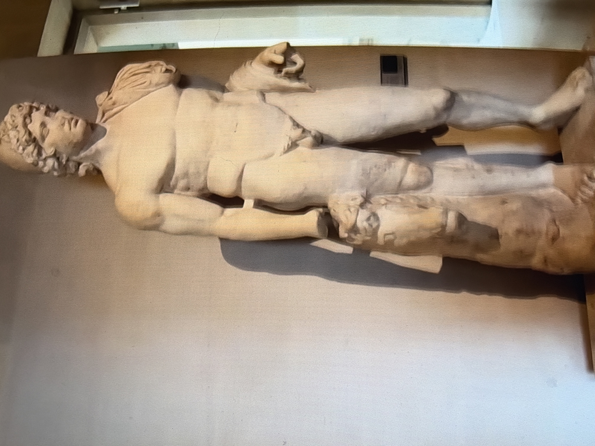 A marble statue of a muscular, nude male figure stands against a light-colored wall. The statue is positioned in profile, facing right, with its left leg forward and its right leg slightly back. The figure has curly hair and wears a helmet or cap. A draped cloth is slung over its right shoulder and hangs down its back, and another cloth is draped over its left arm. The statue is holding a ram or similar animal by its front legs with its left hand. The ram is positioned at the statue's feet. The statue is standing on a pedestal. A shadow of the statue is cast on the wall behind it. To the right of the statue, a vertical element, possibly a doorframe or window, is visible. A dark rectangular object is mounted on the wall behind the statue's right leg. The image is taken from a slightly low angle, looking up at the statue.