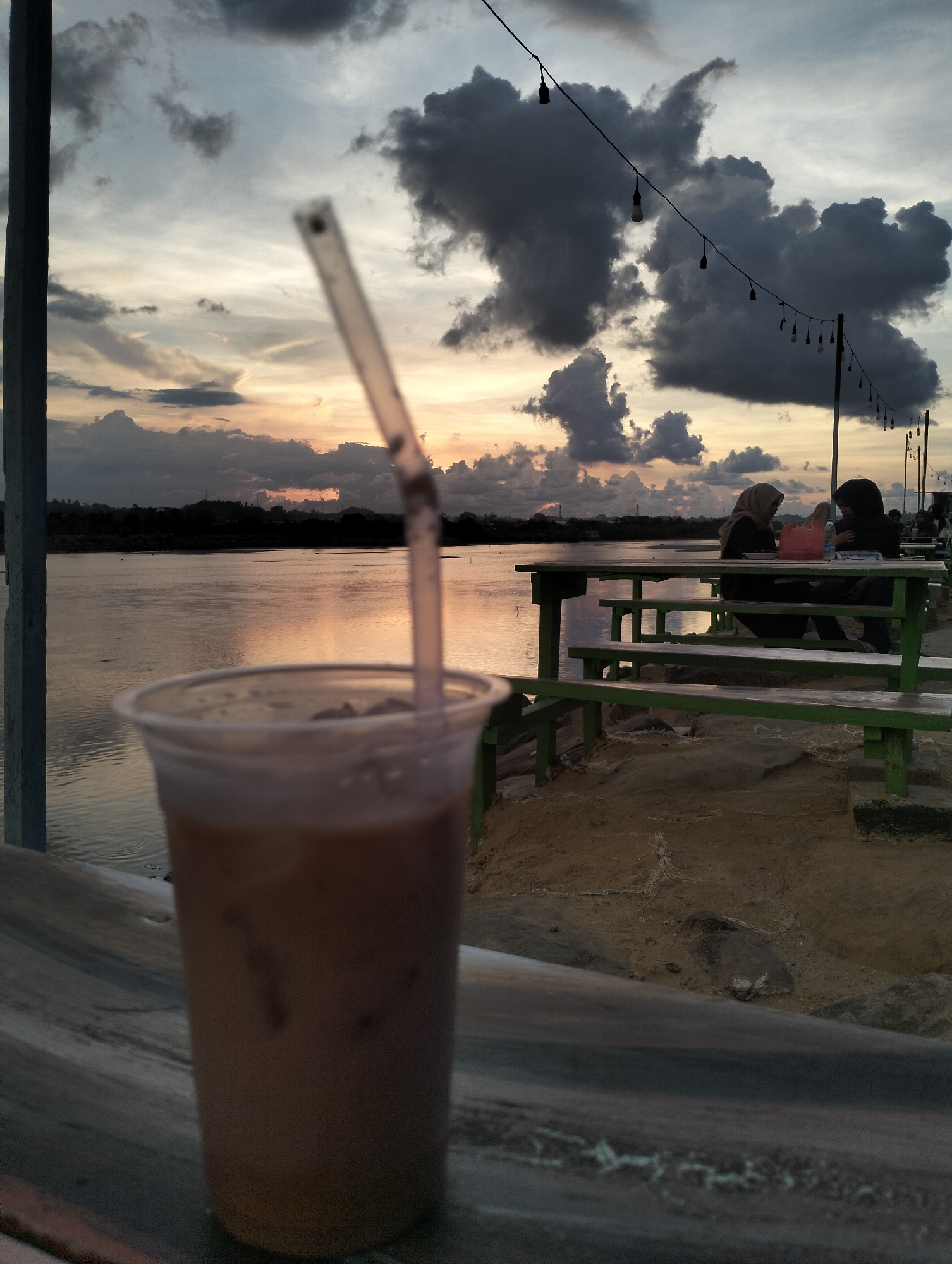 The image captures a scene at what appears to be an outdoor cafe located by a body of water, likely in or near Lhokseumawe, Indonesia. A clear plastic cup filled with a light brown liquid, likely iced coffee, with a plastic straw is in the foreground. A wooden railing is beneath the cup, adding perspective.
In the middle ground, two women wearing hijabs are seated at a green wooden table, which has benches attached to the sides. The table is located on a sandy or rocky area near the water's edge. Behind the table, the water reflects the colors of the sky.
In the background, the sky is filled with dark, dramatic clouds. A string of lights hangs above the tables, suggesting this is a common gathering place during the evening. The distant shoreline has a hazy appearance.