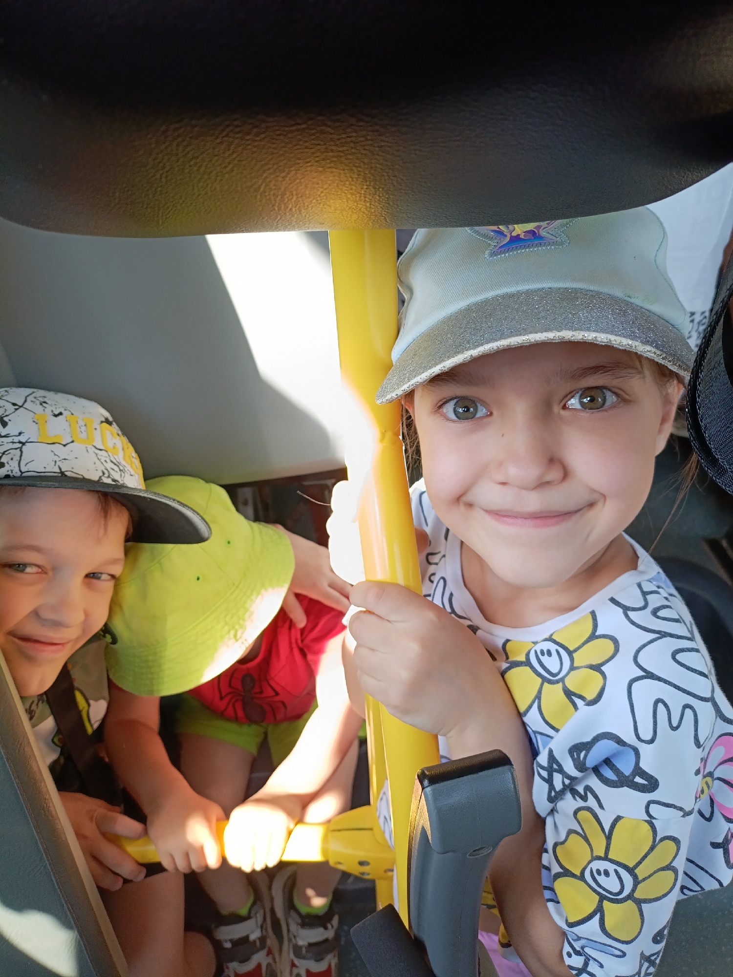 Three children are seated in a vehicle, likely a small bus or cart. The foremost child is a young girl with fair skin, wearing a light-colored baseball cap with a glittery brim, a white shirt with floral patterns, and she is holding a yellow handle. To the left, partially obscured, a young boy with fair skin and a baseball cap is visible, smiling. Behind them, another child wears a bright green hat and a red shirt. The background is a mix of light and dark, with interior surfaces of the vehicle visible. The setting appears to be indoors due to the lighting and vehicle type, though a specific location in Chaikovsky, Russia is not evident from the visual information provided.