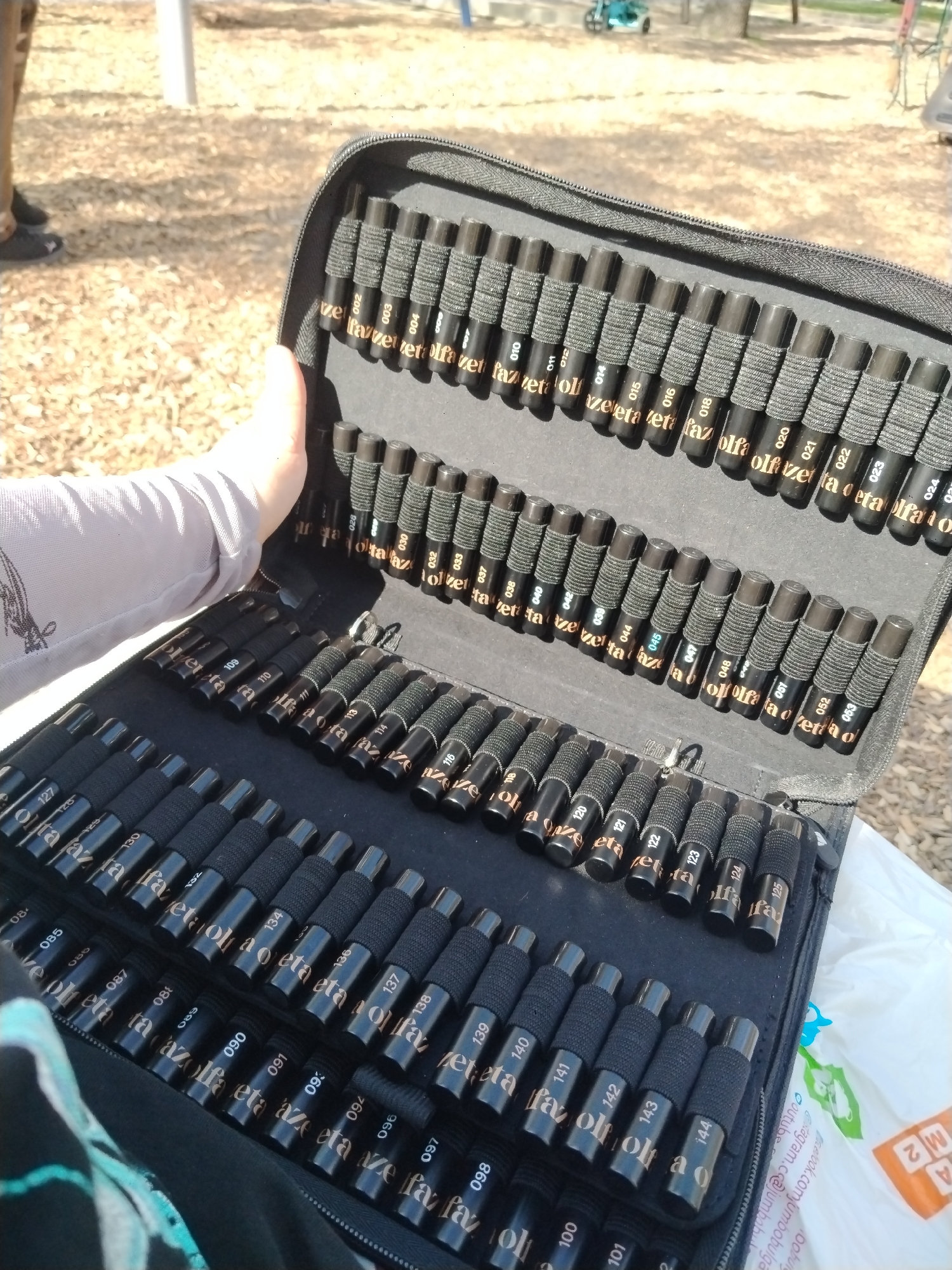 A person holds open a large, black zippered case filled with numerous black markers or pens. The case is designed to hold the markers upright in elastic loops, organized in several rows. Each marker is black with gold-colored numbering and text. The brand name "fazeta olfa" is repeatedly visible on many markers, along with various numerical identifiers such as 002, 003, 004, 010-012, 014-016, 018, 020-024, 028, 030, 032, 033, 037-040, 042, 044, 045, 047, 048, 051-053, 080, 085, 087, 089-091, 093, 096-098, 100, 101, 109, 110, 113, 114, 116, 118, 120-125, 127, 128, 130, 132, 134, 136-144.
The scene is outdoors on a bright, sunny day, indicated by the strong light and clear visibility. The setting appears to be a park or playground in Plovdiv, Bulgaria, with light brown, granular ground cover, possibly sand or wood chips. In the background, parts of what look like playground equipment, including white metal poles, are visible. A second person's legs and feet, clad in dark shoes, are partially seen in the far left background. A light blue or turquoise stroller or tricycle is also visible further back. In the bottom right corner, a white plastic bag is present, featuring logos and text including "youtube", "instagram.cc@jumbobul", "facebook.com/jumbobulga", and "M2".