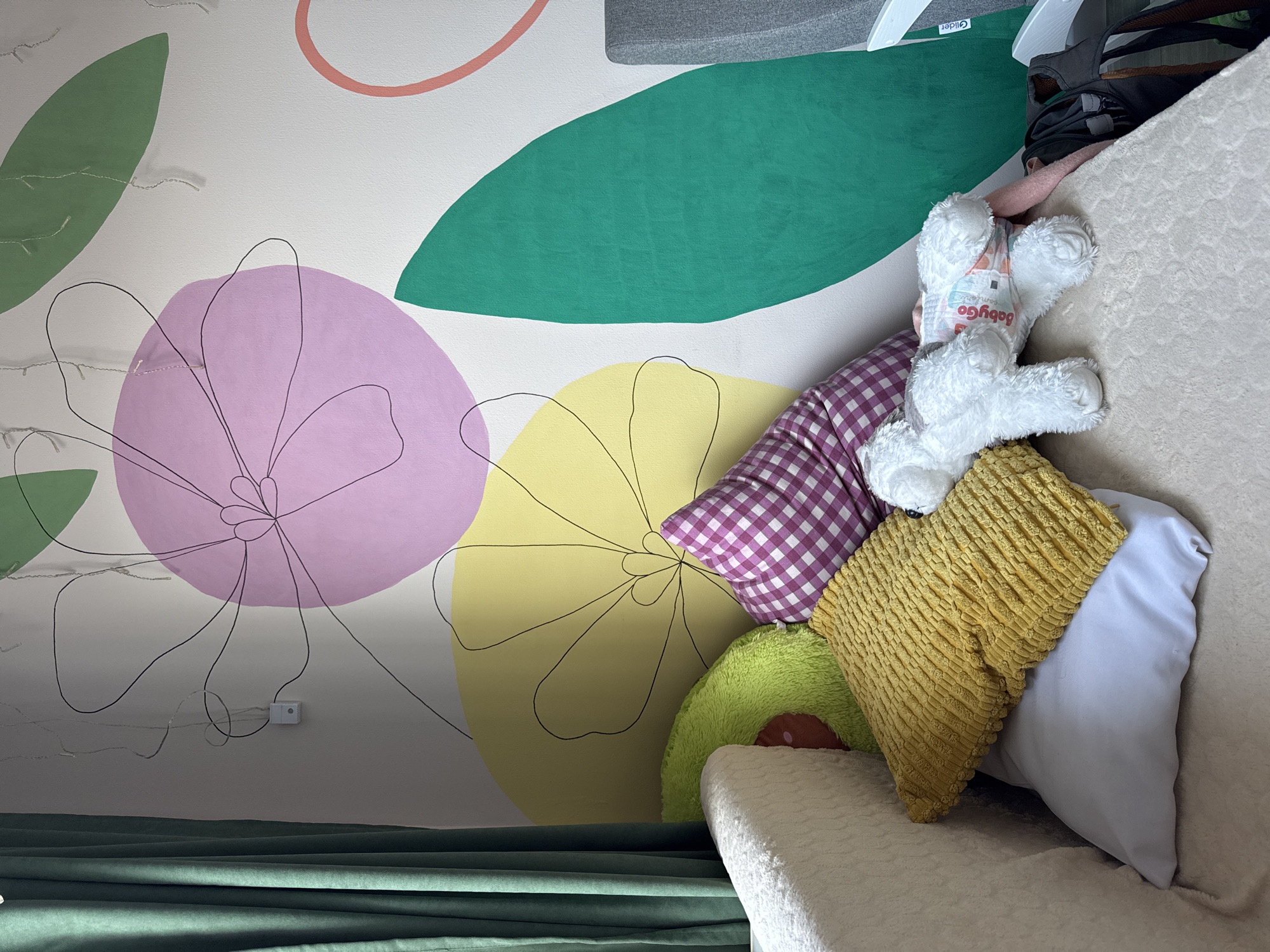 The image shows a section of a room decorated with a playful floral mural on the wall. The mural features large, abstract flowers in colors like pink, yellow, and green, outlined with black lines. There are also string lights with small bulbs running along the wall near the mural.
In the foreground, there are several pillows arranged on what appears to be a couch or bed. A white stuffed animal, possibly a bear with a diaper, is propped up among the pillows. The pillows vary in color and texture, including a purple and white checkered pillow, a mustard yellow textured pillow, a light blue pillow, and a green avocado-shaped pillow. Part of a green curtain is visible at the bottom of the frame.
The setting seems to be a cozy, child-friendly room. The presence of the "Glider" label indicates a potential connection with glider-type seating, but the image does not provide further context. It is not possible to determine if this image was taken in Izhevsk, Russia, without additional information.
