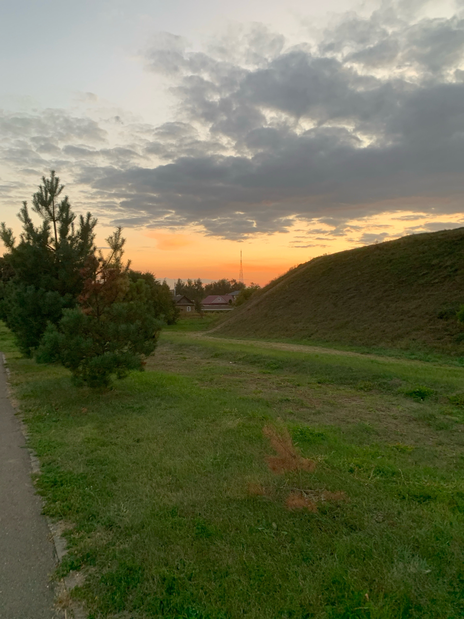 The image depicts a natural landscape at dusk, likely on the outskirts of the city of Kapyĺ, Belarus. In the foreground, a paved path runs along the left side of the frame, bordered by a strip of green grass. The grass continues into the midground, where a cluster of pine trees stands prominently on the left. Further into the midground, a gentle grassy slope rises to the right, forming a significant landform. In the distance, a few houses with red roofs are visible, nestled amongst trees, suggesting a rural or semi-rural settlement. A tall, slender radio tower stands to the left of the houses, silhouetted against the horizon. The sky is filled with a mix of gray and orange clouds, indicating the setting sun. The overall impression is one of a tranquil evening scene in a natural setting.