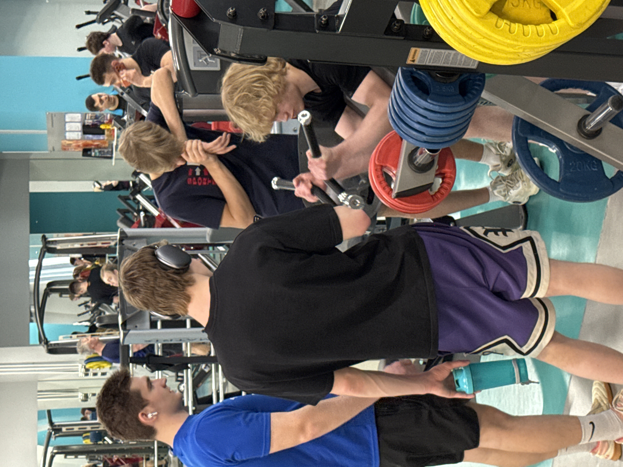 The image depicts a gym setting with multiple individuals engaged in exercise. Several people are using weight machines. The equipment is loaded with various colored weight plates. The gym has a teal-colored floor. The image also shows reflections in mirrors on the walls. The people in the foreground are wearing gym clothes. One is wearing headphones, another a blue shirt and black shorts. The location is not discernible from the image; there is no evidence of Yekaterinburg, Russia.