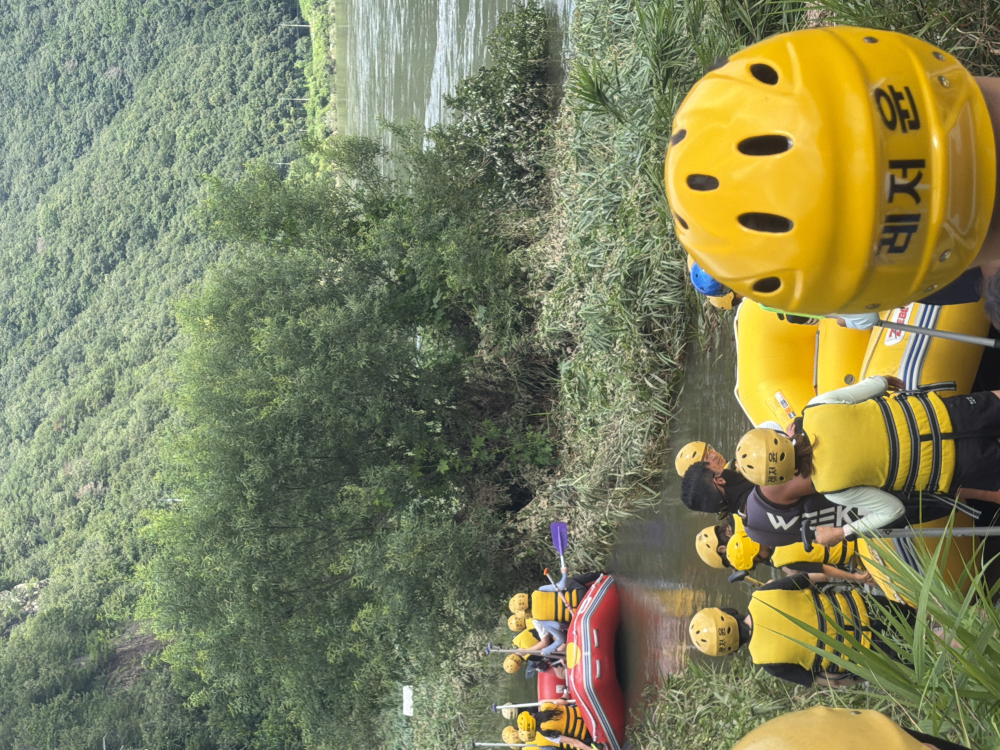 In Seo-myeon, South Korea, a group of people are preparing for a rafting excursion on a river. The scene features lush green trees and a wide, calm river. In the foreground, several individuals are wearing yellow life jackets and helmets, some with Korean text that reads "레저 운" (Leisure Driving). Two red and yellow rafts are partially visible, positioned in the water, suggesting readiness for the activity. One person in the foreground is holding a paddle. The overall atmosphere appears to be one of anticipation for outdoor recreation.