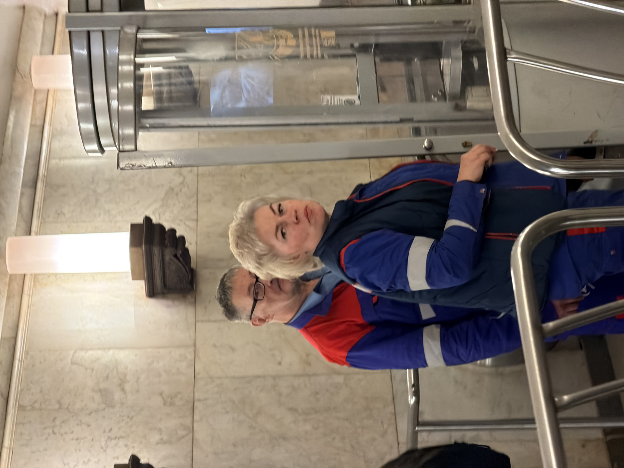 Two people wearing blue and red uniforms are in a building in Moscow, Russia. The woman has blonde hair and is standing next to a man wearing glasses with gray hair. They are standing near a revolving door and a railing. The walls are marble. There is a light fixture attached to the wall.