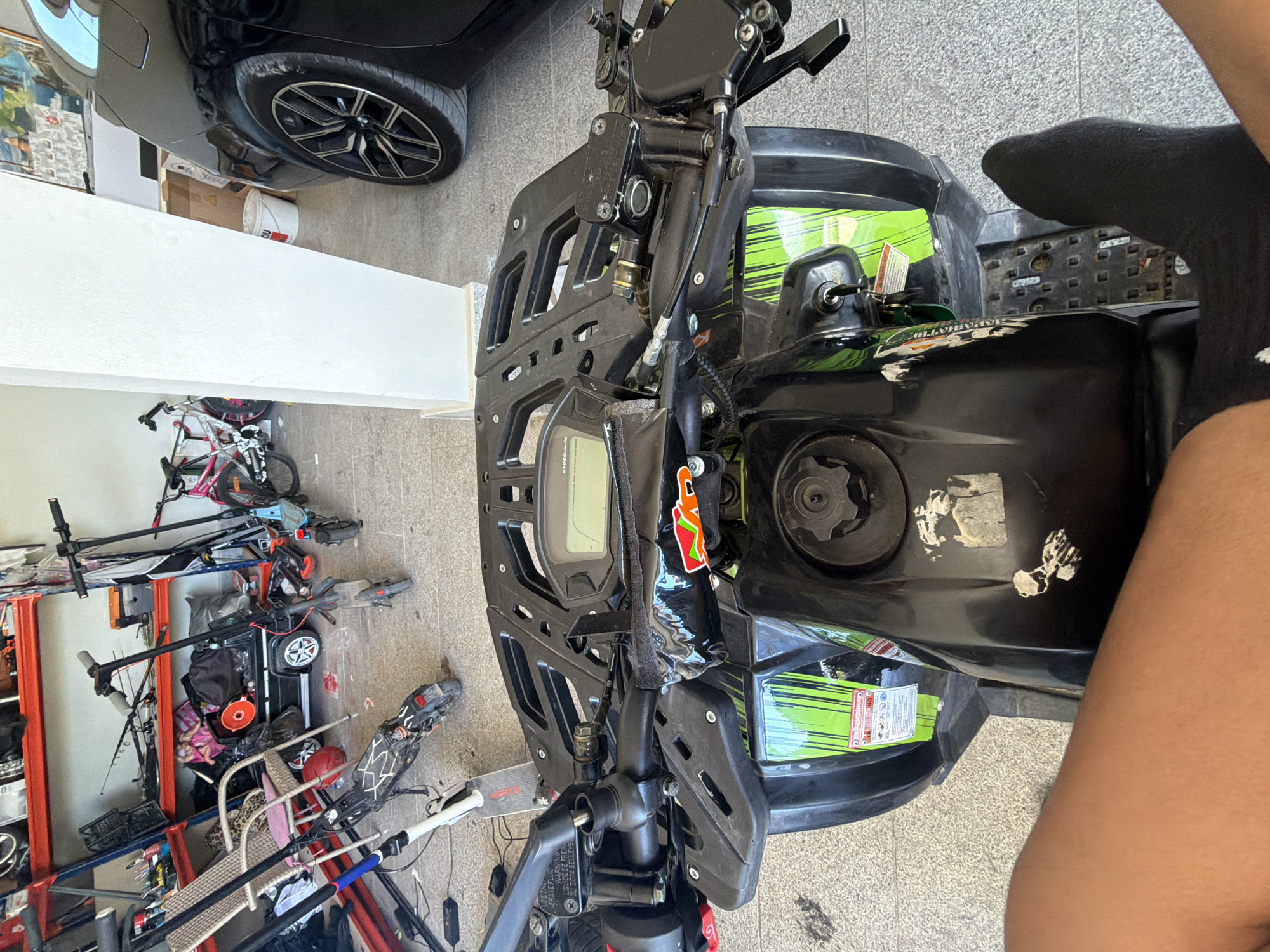 The image shows the front of a black and green all-terrain vehicle (ATV) or quad bike. A black car is partially visible in the upper left corner. Several bicycles, scooters, and other miscellaneous items are arranged against a wall in the background. The ATV is parked on a tiled surface. A person's leg, covered in a black sock, is visible on the right side of the image. There is no information present to determine the location in Asenovgrad, Bulgaria.