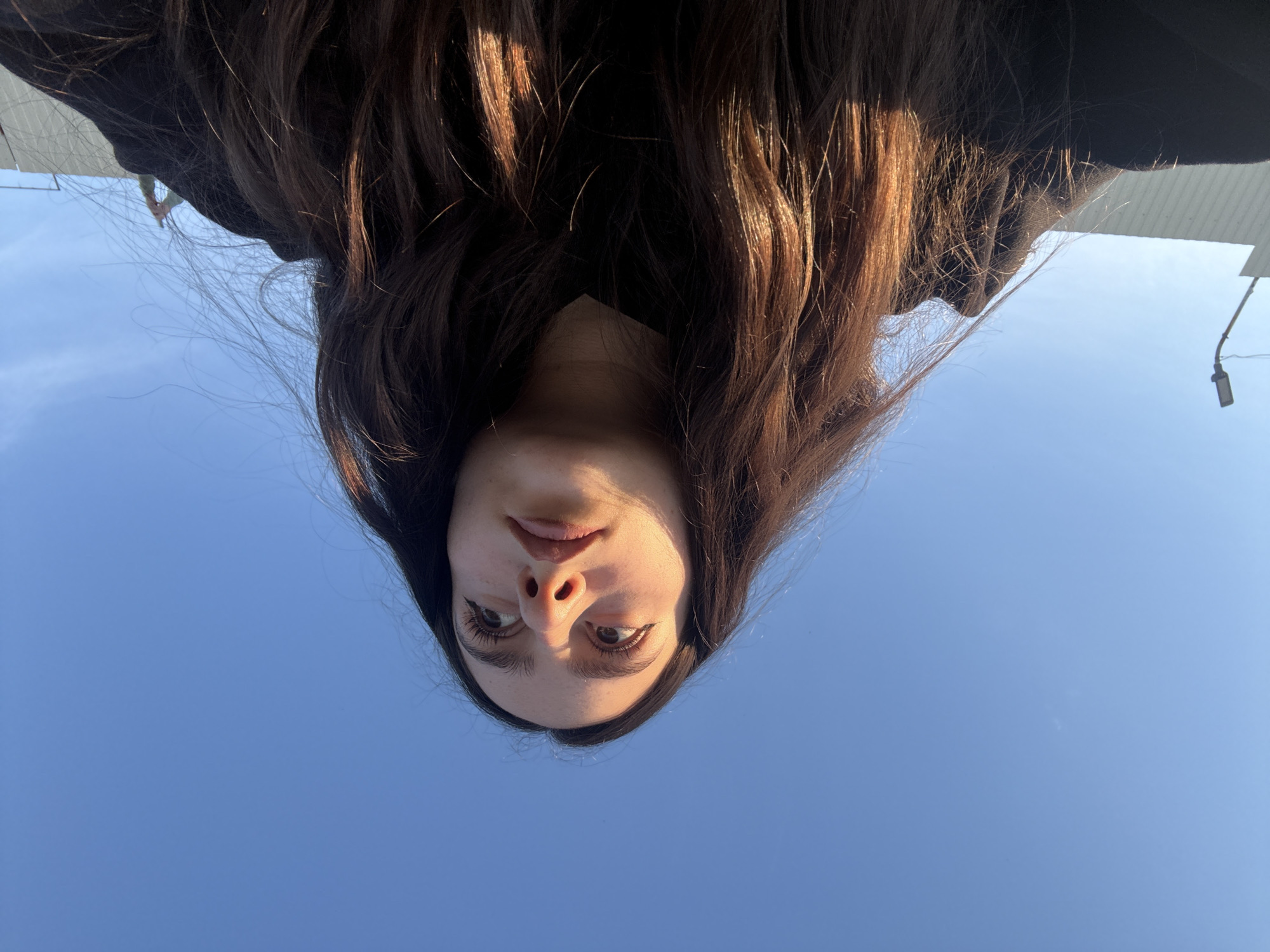 An inverted close-up portrait features a young individual, likely female, captured from a low-angle perspective against a clear blue sky. The subject's head is tilted forward, presenting her face upside down to the viewer. She has long, wavy, dark brown hair with visible lighter brown or auburn highlights that cascade around her face, reflecting sunlight. Her eyes are open, with prominent eyelashes and defined eyebrows, gazing directly towards the camera. Her lips are slightly parted, and her expression is neutral and contemplative.
The dominant background is a vast, unblemished blue sky, indicating bright, clear weather during the daytime, likely mid-morning or afternoon due to the strong, natural lighting. In the periphery, subtle hints of an urban outdoor setting are visible: a light-colored structural edge (possibly a building or awning) appears on the left side of the frame (bottom left in the inverted view), and a dark utility pole or street light fixture extends from the right side (bottom right in the inverted view). The image appears to be a selfie, taken with the camera pointed upwards from below the subject's chin, then rotated or captured with an inverted orientation. The activity is the taking of a portrait, possibly in Yerevan, Armenia. There is no visible text.