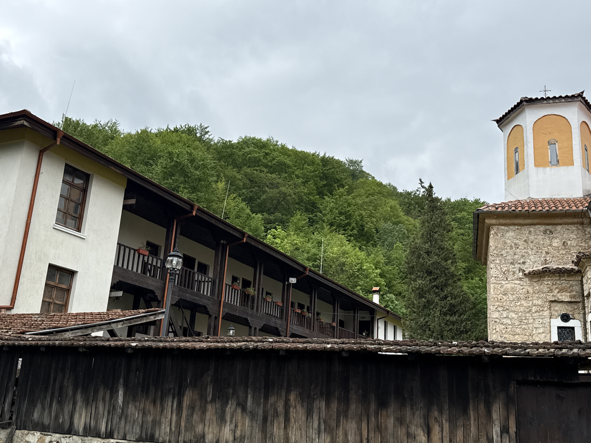 The image depicts the courtyard of the Sveti Georgi Pobedonosets Monastery, located in Ribaritsa, Bulgaria. A long, two-story white building with dark brown wooden balconies and a tiled roof stretches across the left side of the frame. In the foreground is a dark wooden fence or wall. Behind the building is a hillside covered in dense green trees. To the right is a stone building with a bell tower painted white and yellow. The sky is overcast and grey. There are no people visible in the image.