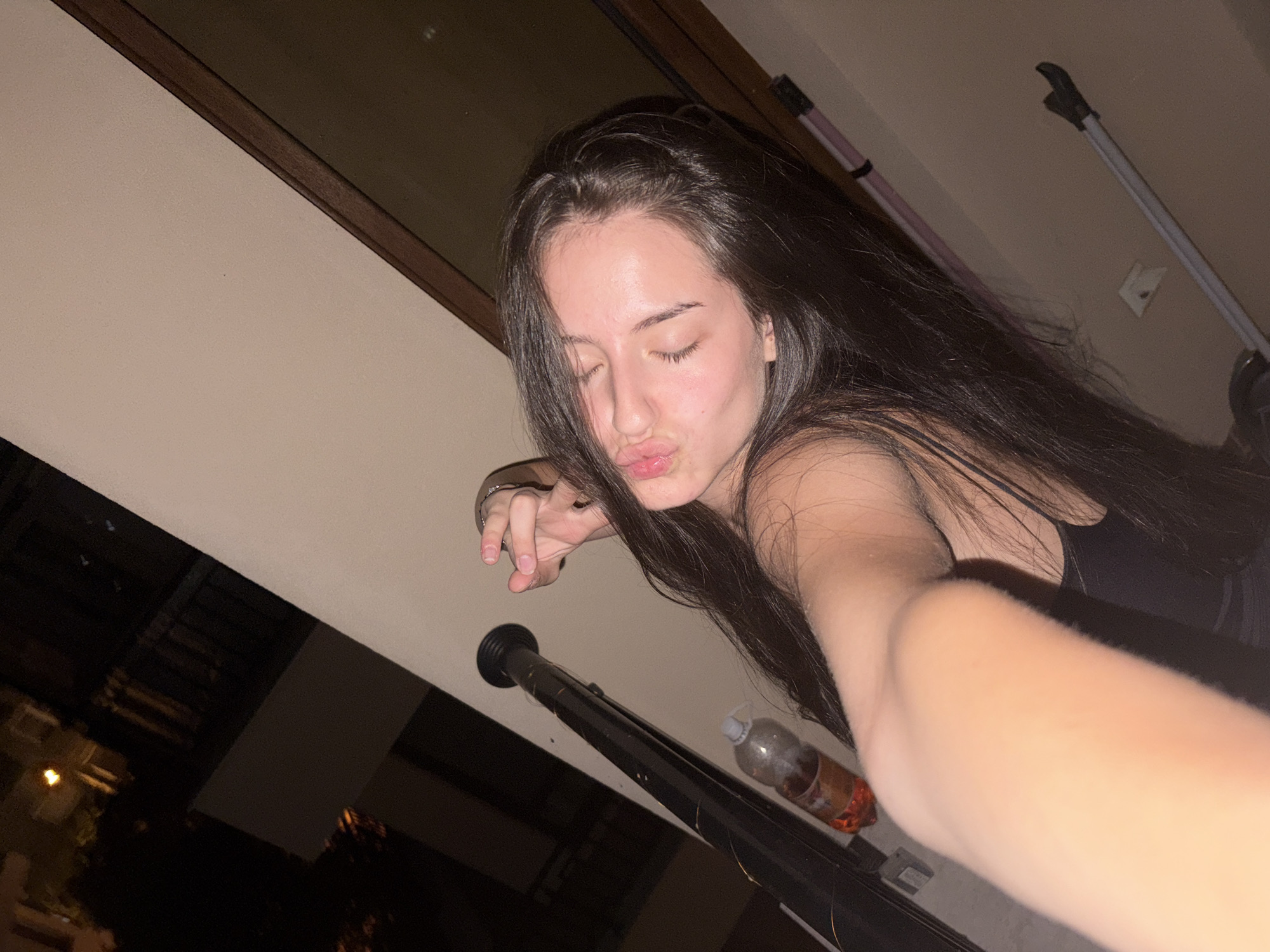 The image shows a young woman taking a selfie with her eyes closed and lips puckered. She has long, dark hair and is wearing a black top. Her right arm is extended, holding what appears to be a smartphone. To her left, a black pole with a silver cylindrical object at the top is visible, along with a clear plastic bottle containing an orange liquid. The background suggests an outdoor setting, possibly a balcony or rooftop at night, with faint lights from a building visible in the distance. The scene is likely located in the city of Burgas, Bulgaria, given the context provided.