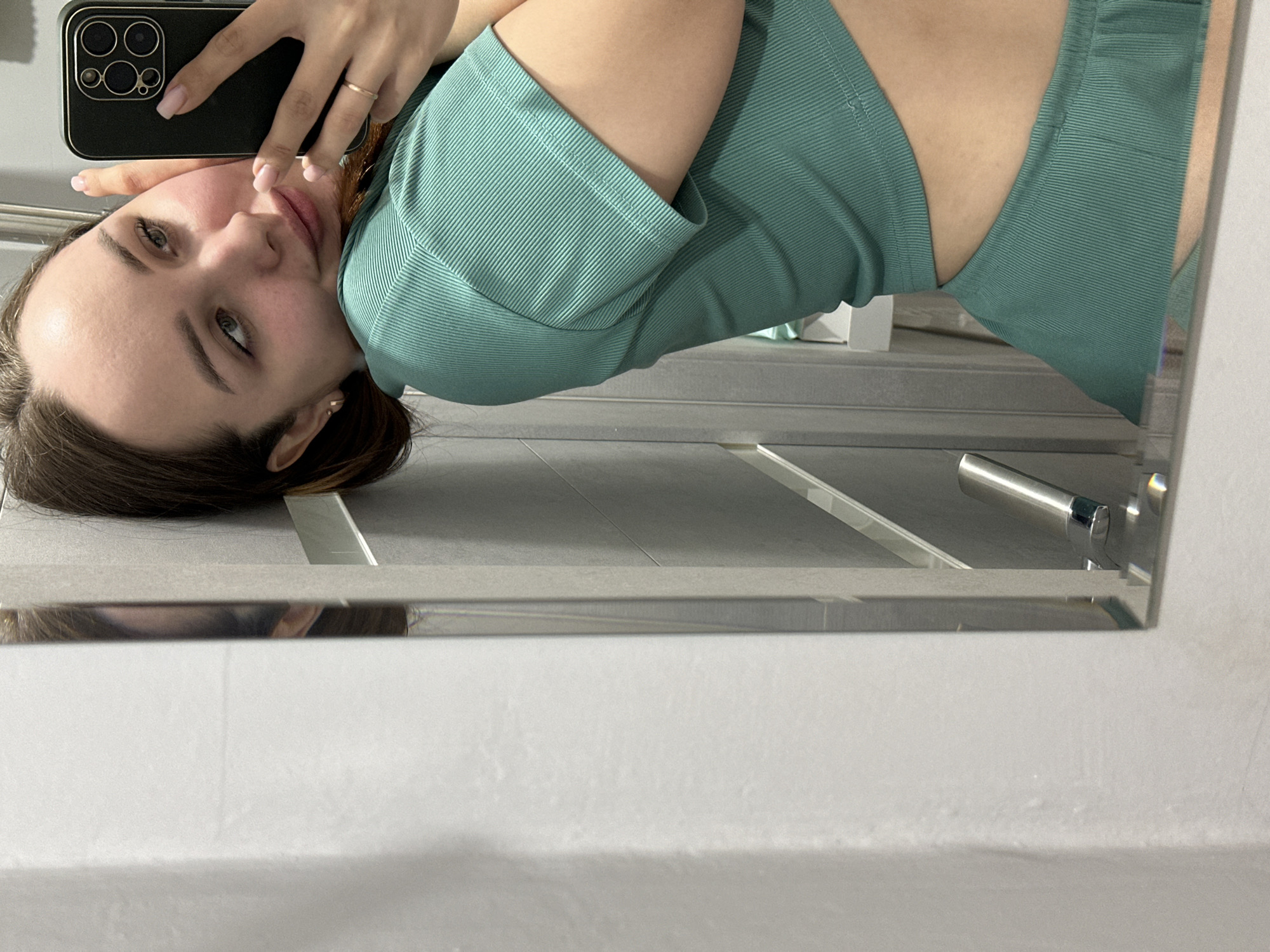 A young woman, appearing to be in her late teens or early twenties, is captured in a mirror selfie, oriented sideways in the frame. She has light brown hair and light eyes, and her complexion is fair. She wears a seafoam green or teal ribbed short-sleeved top and matching bottom, possibly shorts or pants. A subtle, serene expression is on her face as she looks towards the camera's reflection. Her nails are painted a light pink, and she wears a simple gold ring on her right ring finger.
She holds a dark-colored smartphone, likely an iPhone due to the distinct camera cluster on its back, to take the photo. The phone is held in her left hand, positioned near her face. The selfie is taken indoors, likely in a bathroom or dressing room in Chaikovsky, Russia, as suggested by the tiled floor visible in the reflection and a metallic fixture, possibly a door handle or towel rail, also reflected in the mirror. The lighting appears to be ambient indoor light. The background is minimalist, with light-colored walls and the reflective surface of the mirror occupying a significant portion of the image. No visible text is present.