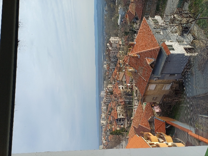 The image displays an elevated view of a residential area in Gorna Oryahovitsa, Bulgaria, seen through a window frame. The top portion of a dark, dusty window frame and a segment of a light-colored vertical frame on the left are visible. The sky is partly cloudy, featuring light blue patches and scattered white stratocumulus clouds.
In the foreground, on the left, a multi-story building with a yellow-orange facade and curved balconies is present. Below it, a paved walkway or narrow street runs parallel to the left frame. To the right, a prominent two-unit or two-section building features distinct terracotta-tiled pitched roofs. One section of this building is painted light grey/white, exhibiting multiple windows and a balcony. The adjacent section, slightly behind, is light brown/beige with a simpler facade. A deciduous tree with bare branches stands in front of the grey section of this building.
The midground reveals a densely packed urban environment, primarily composed of two to three-story houses with characteristic orange terracotta roofs. Several taller, multi-story apartment blocks, typically lighter in color and rectangular, are interspersed among the houses. Streets, bare trees, and utility poles with overhead lines are visible throughout this residential expanse.
The background extends into a broader plain, where the city gradually diminishes towards a distant, low-lying horizon. A faint outline of hills or mountains is discernible on the far horizon. No discernible individuals or moving vehicles are visible within the depicted scene.