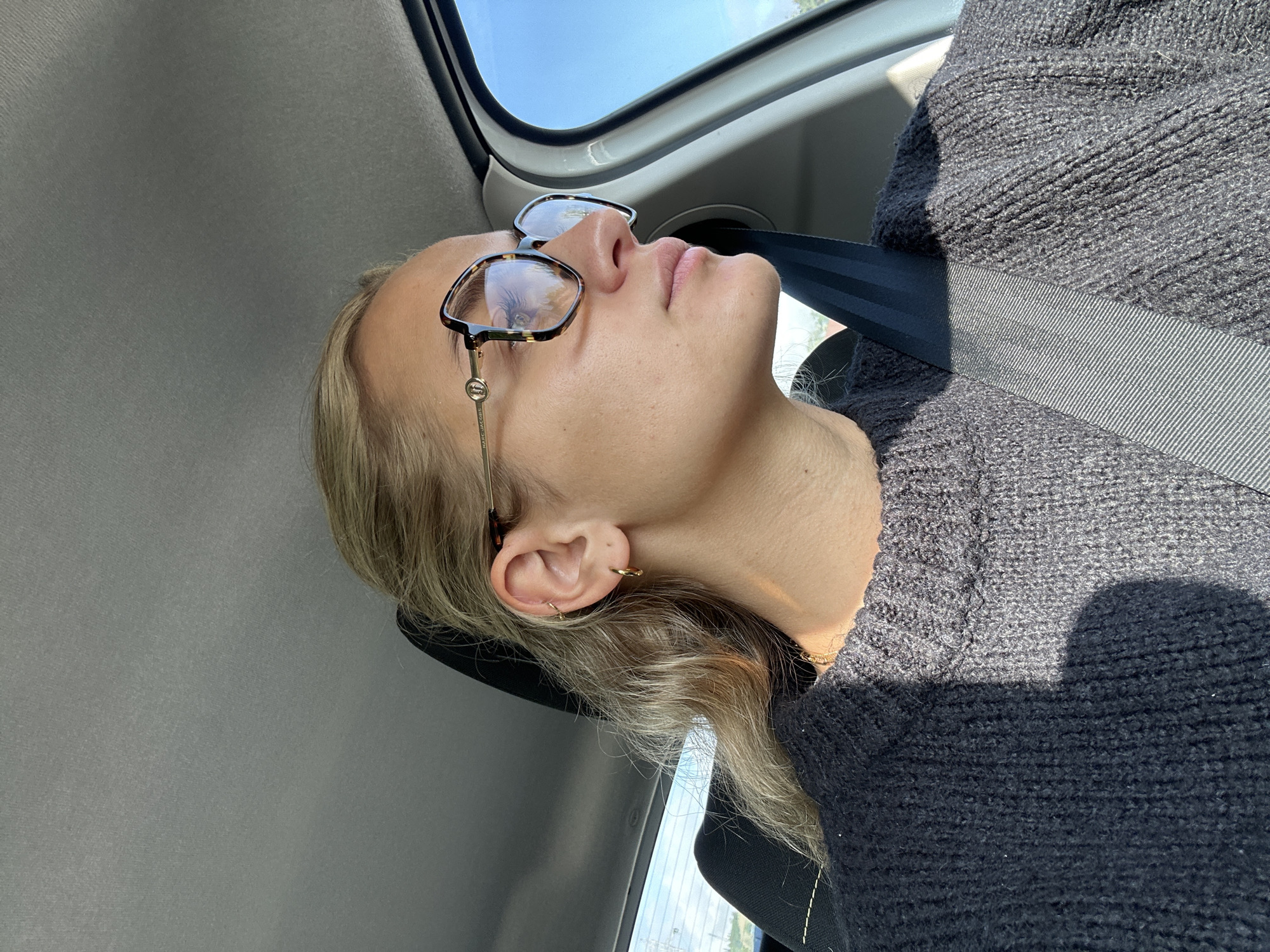 A young person with blond hair is shown resting or sleeping in the passenger seat of a car. Her head is tilted back, with eyes closed or downcast, and her mouth slightly agape. She wears a dark, chunky knit sweater and a grey/black seatbelt secured across her body. On her face are tortoiseshell-framed glasses with thin gold temples, clearly displaying the brand "MARC JACOBS" on the arm. Small gold hoop earrings adorn her left ear.
The scene is set inside a vehicle, with a light-colored ceiling and window frames visible. Through the car windows, a clear blue sky and glimpses of distant scenery are visible, indicating daylight. Strong sunlight illuminates the scene, casting prominent shadows on the person's sweater and face. The overall impression is one of peaceful travel or a momentary rest during a journey in Chabanière, France.