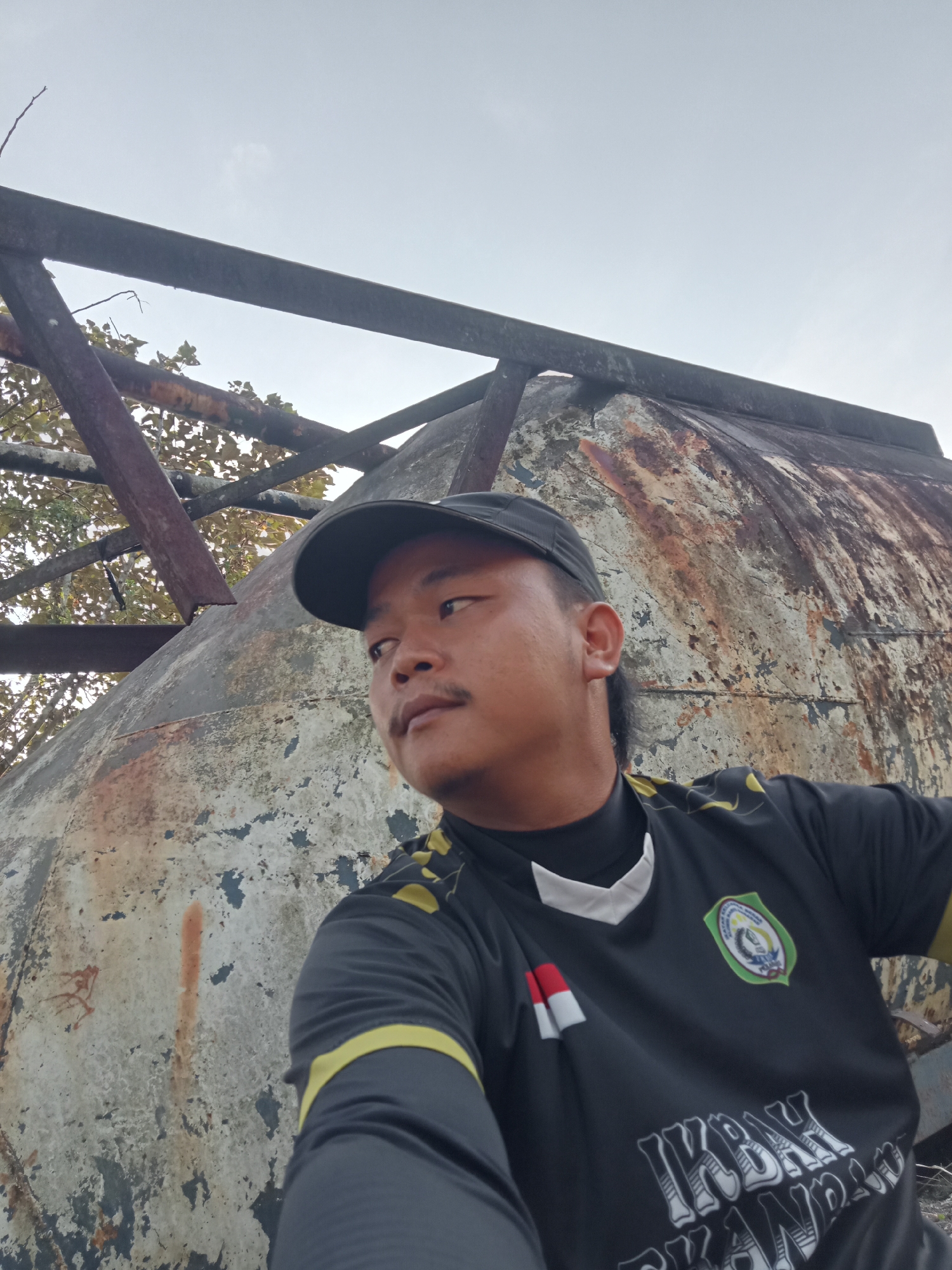 A medium shot from a low angle shows a man in a black t-shirt with yellow accents and a logo on the left chest. He is wearing a black baseball cap and looking to his right, away from the camera. The man has a mustache. Behind him, a large, rusted, curved metal structure dominates the background, with a section of broken metal framing visible above it. Some trees with brown leaves are seen through the framing on the left. The sky is overcast and pale gray. The scene appears to be outdoors, possibly in an industrial or abandoned area.