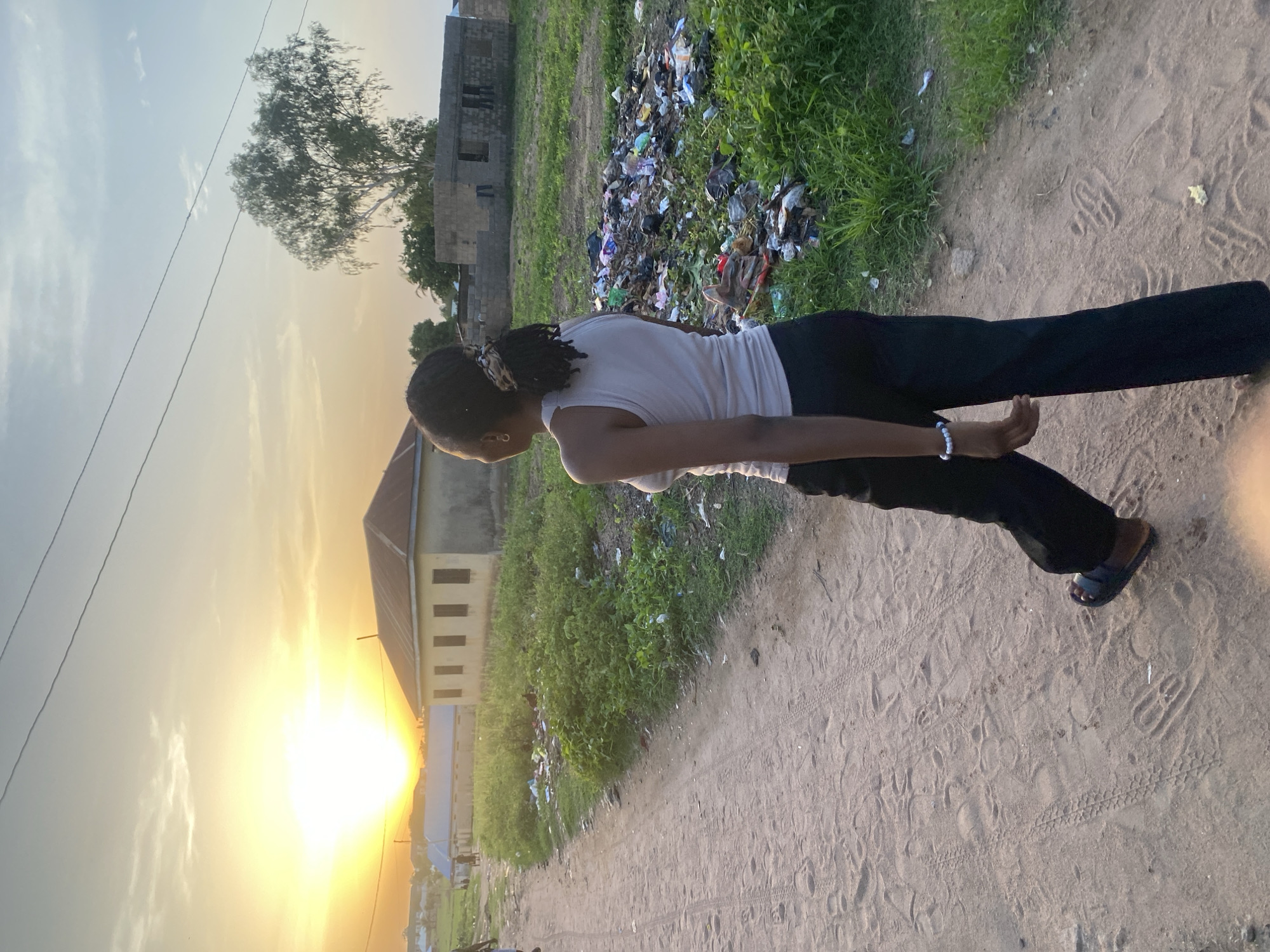 A young Black woman is walking away from the camera on a dirt road in Jauro Yinu, Nigeria. She is wearing a light-colored tank top, black pants, and sandals. Her hair is in braids tied back with a leopard print scrunchie. To her right, there is a large pile of garbage and debris in an overgrown grassy area. Beyond the garbage, there are some unfinished buildings constructed with concrete blocks, and a single tree. Further in the background, a row of houses with metal roofs are visible under a bright, hazy sunset sky. The road has tire tracks and footprints in the sandy soil.
