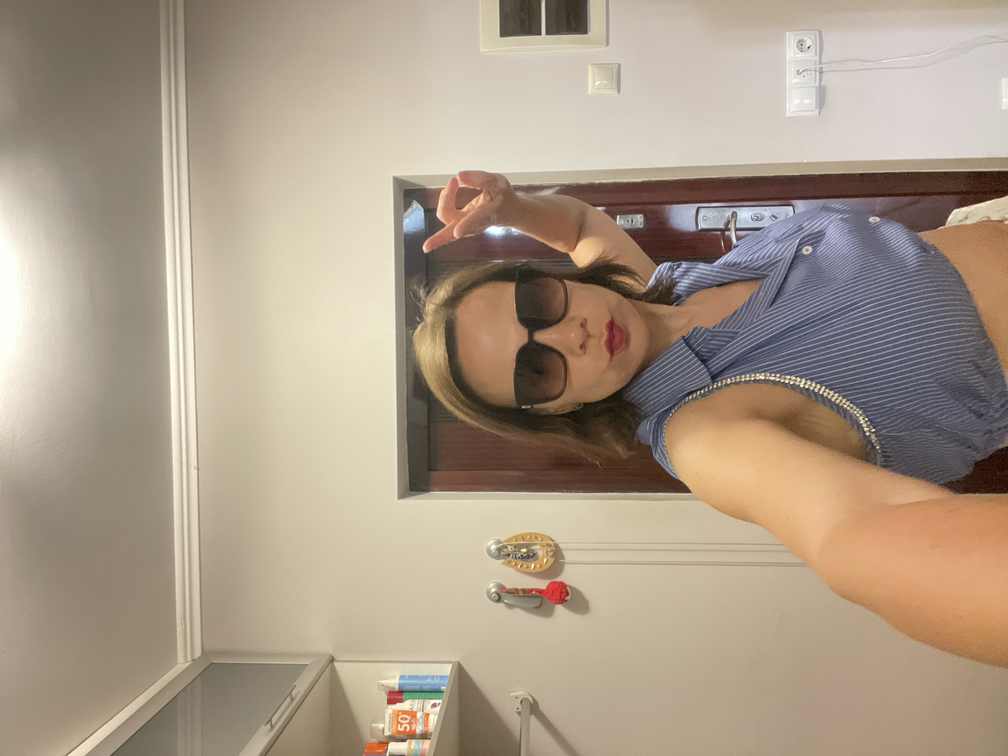 A woman with brown hair and sunglasses is standing in a room, posing for a photo. She is wearing a blue and white striped shirt with decorative detailing on the shoulder straps. Her left arm is raised, and her fingers are in a gesture. Behind her, a dark wooden door is visible, along with a wall with electrical outlets and a white switch. To her left, a shelf unit is mounted on the wall, containing several bottles, one of which is labeled "50". The room appears to be a domestic interior. There is no information in the image that places this scene in Sochi, Russia.