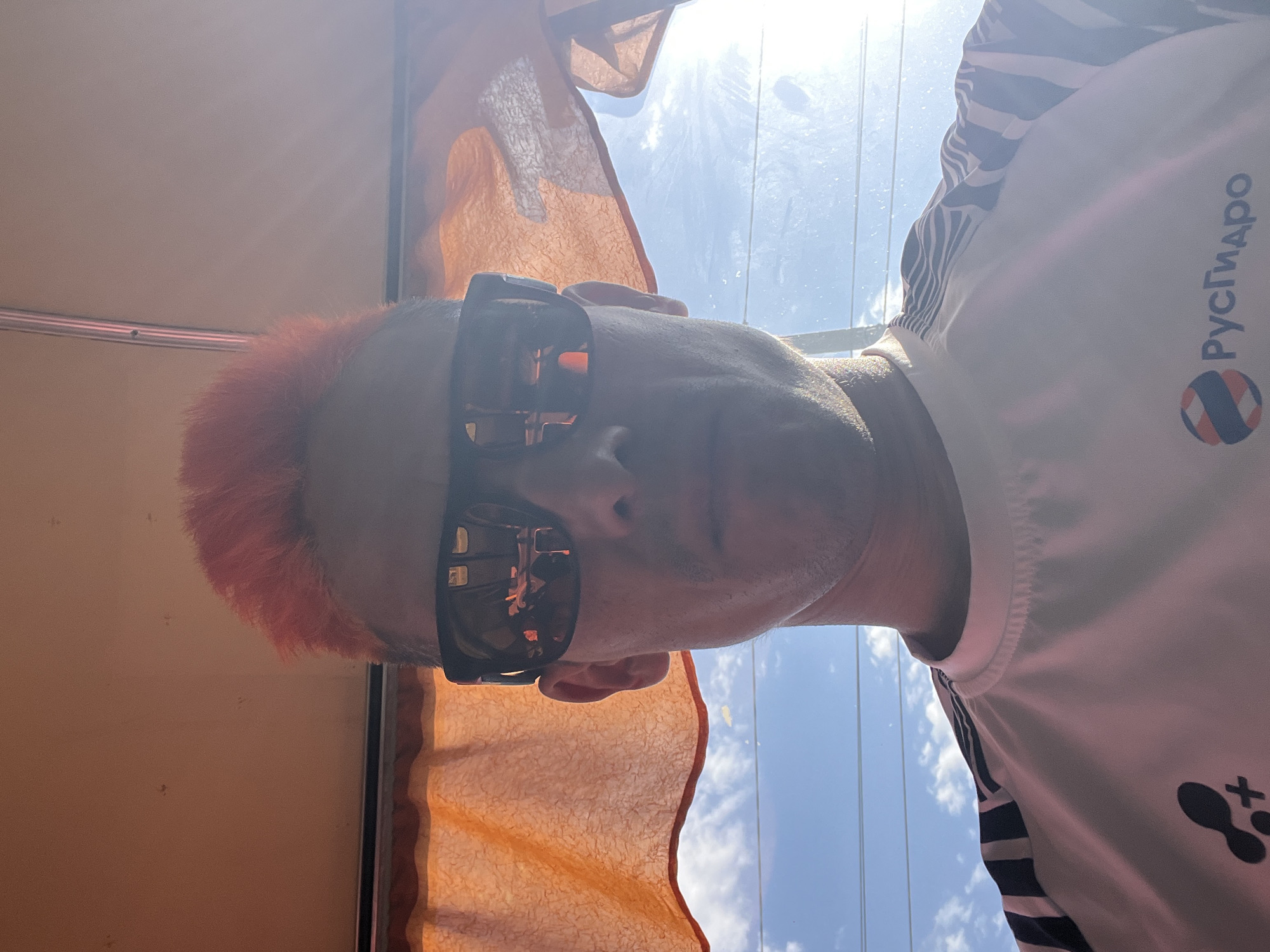 A man wearing sunglasses is positioned near a window. The man has short, orange hair and is wearing a white shirt with black stripes along the sleeves. There is a logo with the word "РусГидро" on the right side of the shirt. Through the window, a bright sky with some thin clouds can be seen. There's an orange curtain or fabric near the window. The photo was taken in Chaikovsky, Russia.
