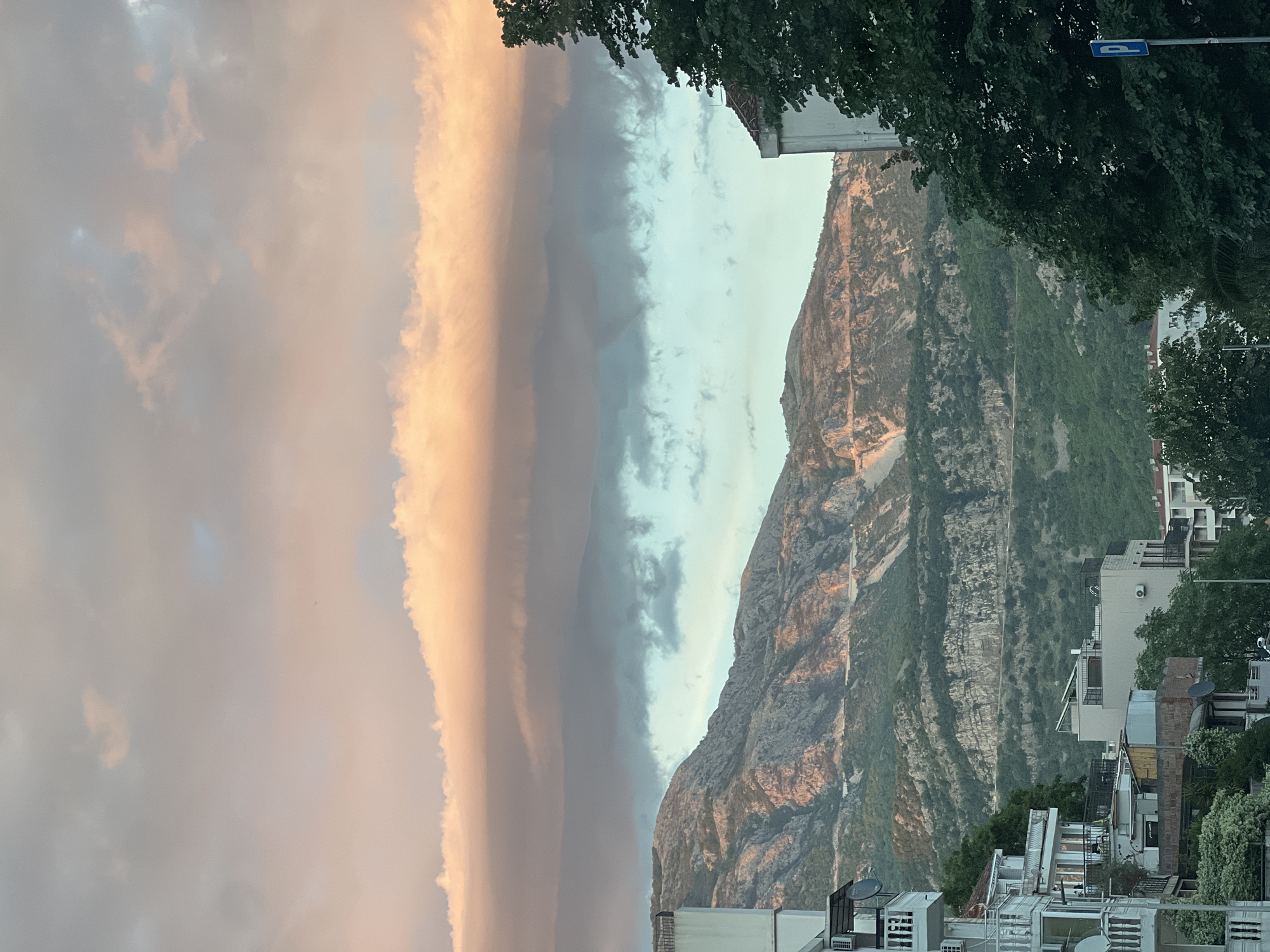 This media file presents a panoramic view of a mountainous landscape under a dramatic sky, characteristic of a coastal town like Budva, Montenegro. No people are visible.
The scene is dominated by a rugged, sprawling mountain range that rises from the mid-ground, featuring rocky slopes interspersed with dense green vegetation. A winding path or road is visible on the mountain's face. In the foreground and extending along the base of the mountain are numerous residential buildings, primarily white or light-colored, with multiple stories, flat roofs, balconies, and architectural details like satellite dishes and air conditioning units. Lush green trees are scattered among the buildings and frame the upper right portion of the image.
The sky is a notable element, displaying a striking contrast of colors and cloud formations. On the left, clouds are illuminated with warm, soft hues of peach, orange, and gold, suggesting either a sunrise or sunset. These blend into darker, cooler-toned grey and pale blue clouds toward the right, creating a dynamic atmospheric effect.
A blue rectangular road sign with an indecipherable white symbol is partially visible through the foliage in the upper right corner. The low-angle light on the mountains and the warm glow on the clouds strongly indicate late afternoon or early evening. The overall atmosphere is serene and scenic, capturing the natural beauty and urban development of the location.