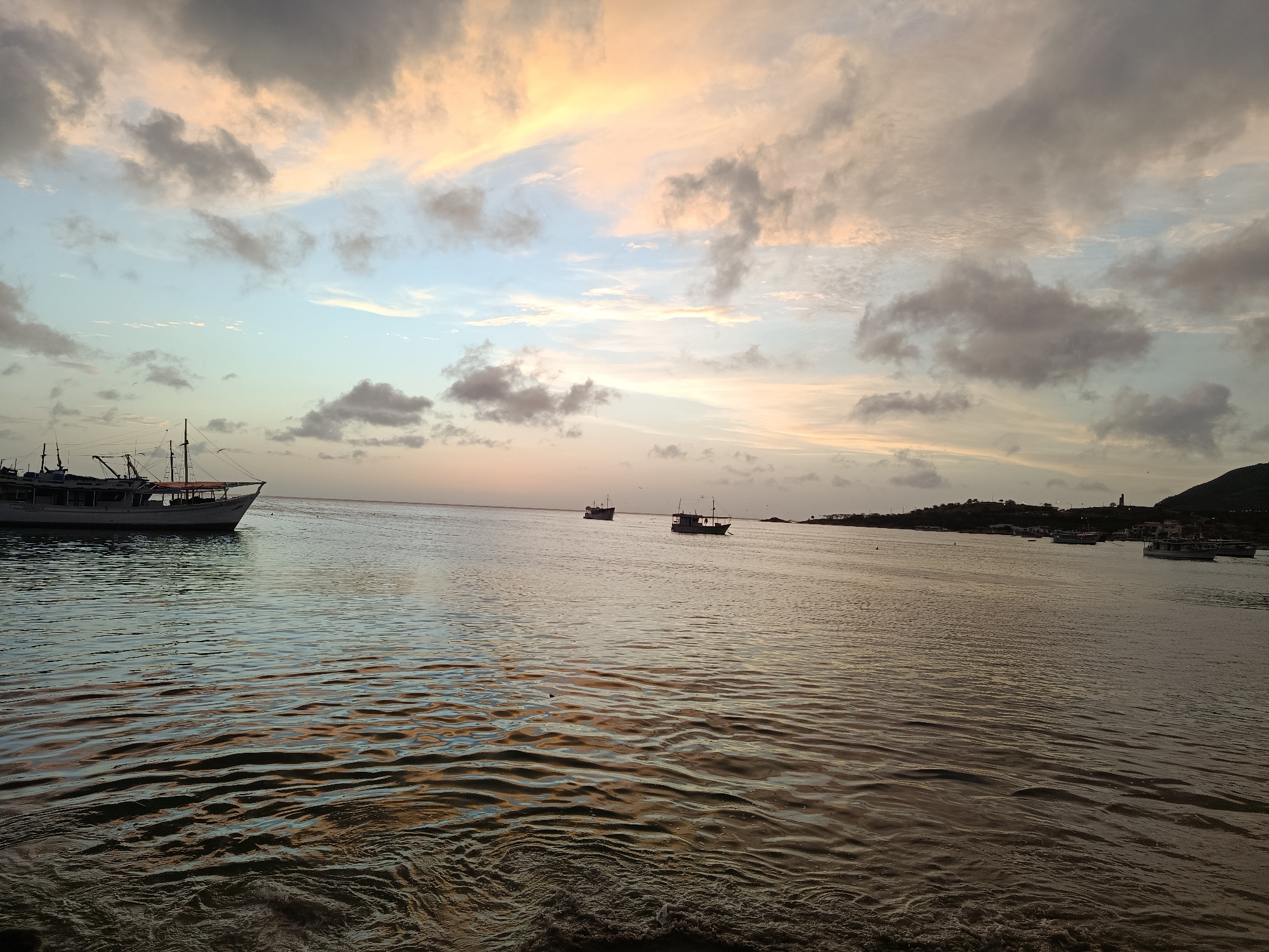 The image depicts a serene coastal scene at Juan Griego, Venezuela, likely during sunset or sunrise. A large white boat with multiple masts and rigging is anchored in the foreground on the left side of the frame. The water is calm with gentle ripples reflecting the warm hues of the sky. In the middle ground, two smaller fishing boats are visible on the horizon. The background features a faint silhouette of the coastline with rolling hills and scattered structures. The sky is a canvas of soft pastel colors, with pale oranges, pinks, and blues dominating the clouds. The overall impression is one of tranquility and natural beauty.