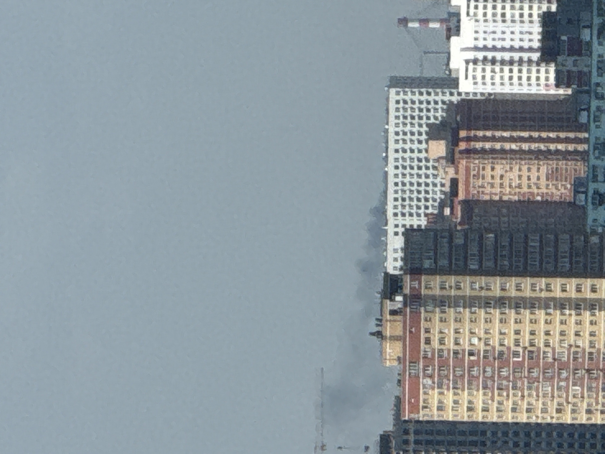 The image shows a cityscape with several tall residential buildings under a hazy, light blue sky. In the background, a thin plume of grey smoke rises from behind a cluster of buildings, partially obscured by a crane. The buildings are modern high-rises with repetitive window patterns. One building in the foreground is particularly prominent, with a facade of alternating yellow and red vertical stripes. The overall scene suggests an urban environment, possibly a construction site or an area affected by an incident causing the smoke. The location is identified as Lyubertsy, Russia.