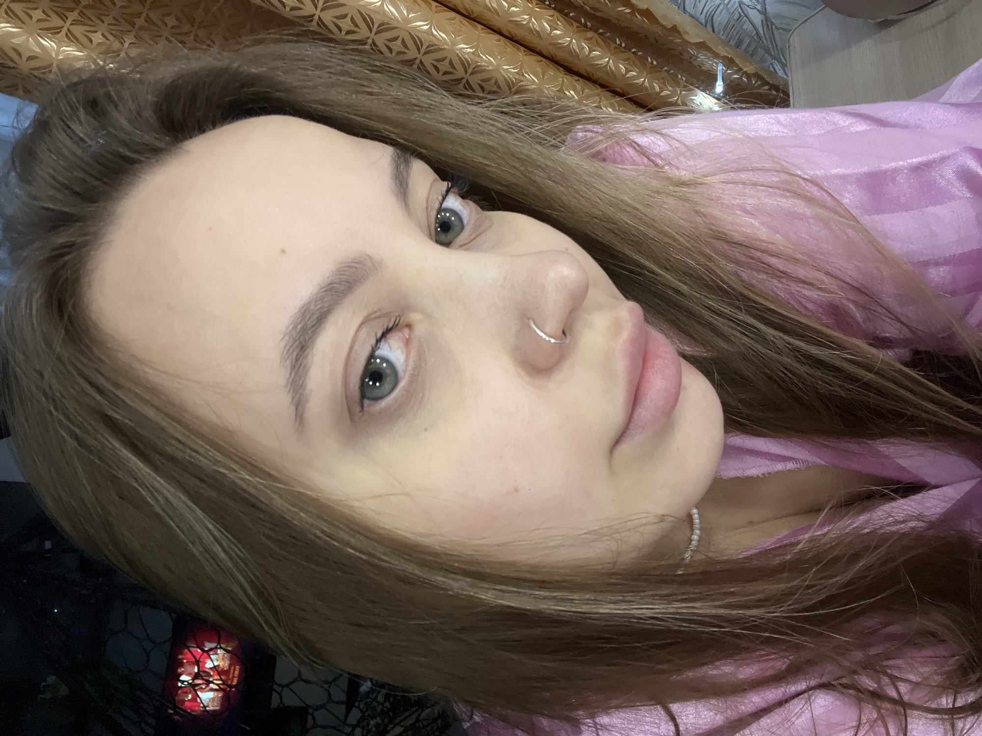 A close-up, tilted selfie captures a young woman with light brown hair and gray-green eyes. She has a silver ring in her left nostril and wears a pearl necklace. She is dressed in a pink, striped garment. The background includes a patterned curtain, an indiscernible object in the lower left corner with a red light, and a partially visible surface. There is no information available in the image to identify the location as Novopokrovskaya, Russia.