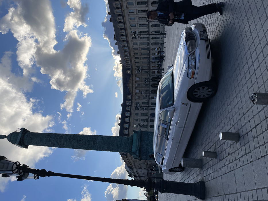 A white stretch limousine is parked on the cobblestone pavement in Place Vendôme, Paris, France, under a partly cloudy sky with visible crepuscular rays emanating from behind clouds in the upper right. The Vendôme Column, a large green bronze column adorned with spiral bas-reliefs and topped with a statue, stands prominently in the background to the left of the image center. Surrounding the square are classical Parisian buildings with numerous windows, mansard roofs, and chimneys, characteristic of the city's architecture.
In the foreground on the left, a tall, ornate black lamppost is visible. Several short, silver-colored cylindrical bollards are embedded in the pavement near the lamppost and in front of the limousine. A male individual, dressed in a dark suit jacket, light blue shirt, and dark trousers, stands on the right side of the image, facing slightly left towards the limousine. He holds a dark object, possibly a camera or smartphone, in his hands. His attention appears directed towards the vehicle or the surrounding area. The limousine's front license plate, "AK-159-PP," is partially visible. Other indistinct figures are visible walking in the distance across the square. The sunlight casts shadows, indicating a low sun angle, suggesting late afternoon or early evening.
