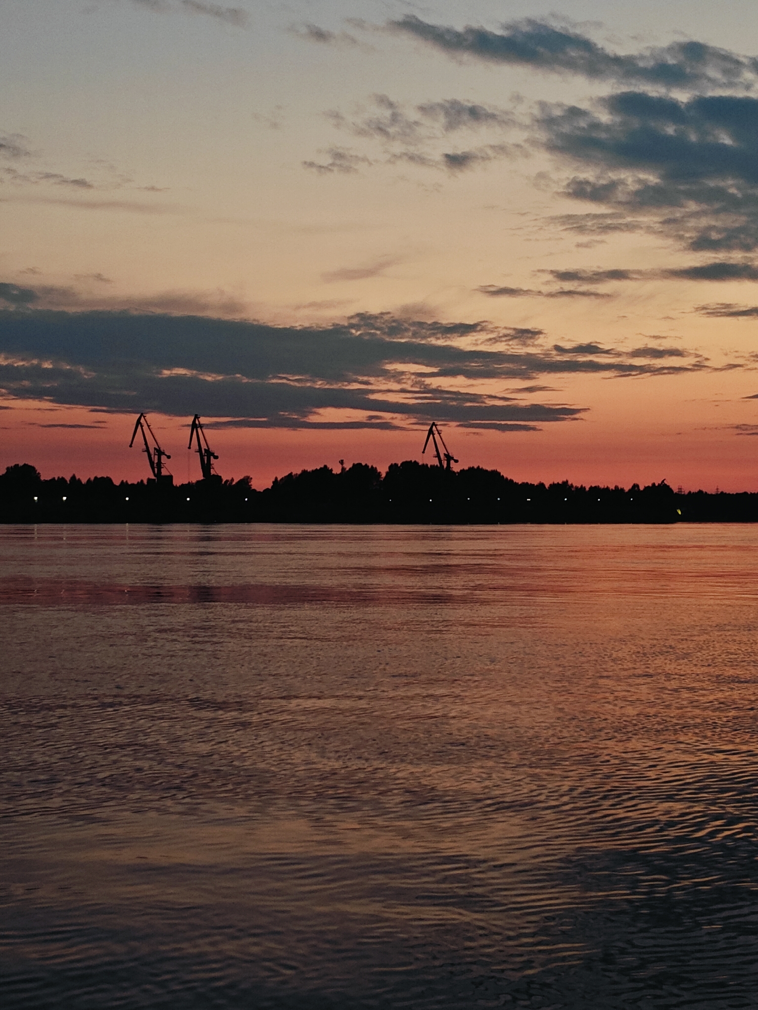 This is a silhouette photograph taken at sunset, likely over the Kama River in Chaikovsky, Russia. The foreground features the calm, rippling water of the river, reflecting the warm hues of the sky. In the midground, a silhouetted treeline marks the bank, punctuated by several industrial cranes. These cranes, along with scattered lights along the shore, suggest a port or industrial area. The sky is a blend of soft oranges, pinks, and purples, with streaks of dark clouds adding texture. The overall mood is tranquil and atmospheric, typical of a sunset scene. No people are visible in the image.