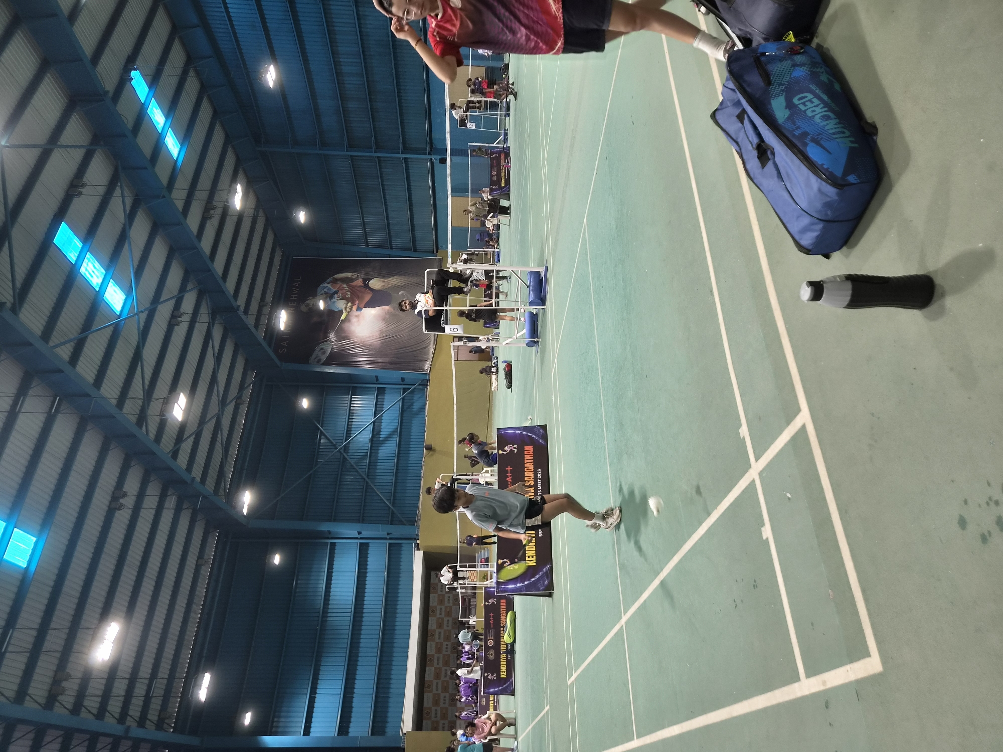 The image depicts an indoor badminton court located in Phagwara, India. The court is illuminated by overhead lights and features a green synthetic floor with white court lines. Several individuals are present on and around the court, participating in or observing a badminton match. In the foreground, a young boy in a grey t-shirt and black shorts is mid-jump, about to hit a shuttlecock. To the right, a person wearing a red and black t-shirt and black shorts is partially visible, with their hand touching their head. Two large blue badminton bags and a black water bottle are placed on the court floor near the foreground. In the background, there are spectators, officials seated on elevated chairs, and other players. A large banner featuring an image of a badminton player hangs on the back wall, along with tournament advertisements. The overall environment suggests an active sporting event taking place within a modern sports facility.